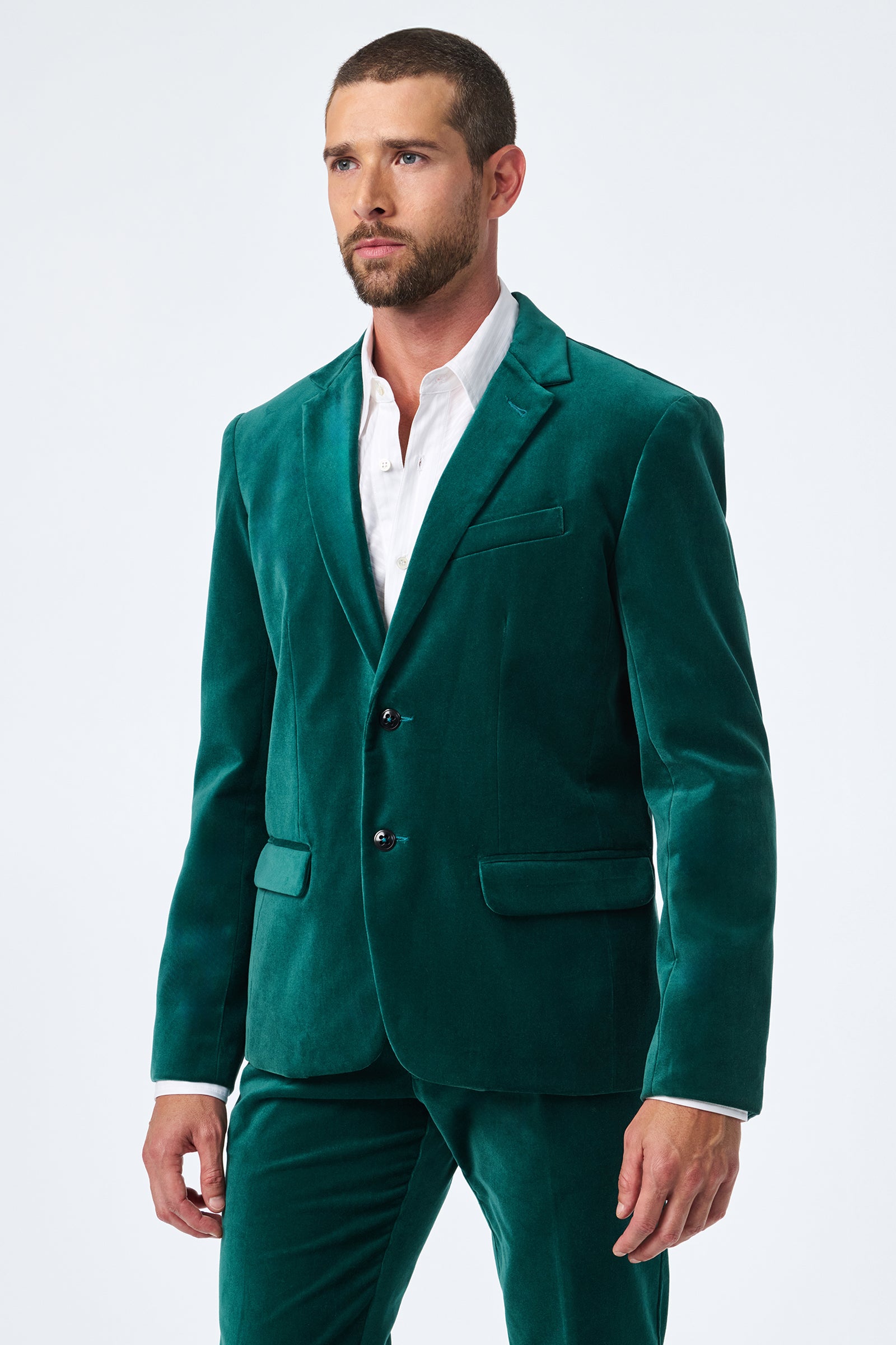 A man with short brown hair and a beard wears the Thurston Velvet Blazer in green with matching pants over a white dress shirt, standing against a plain light background.