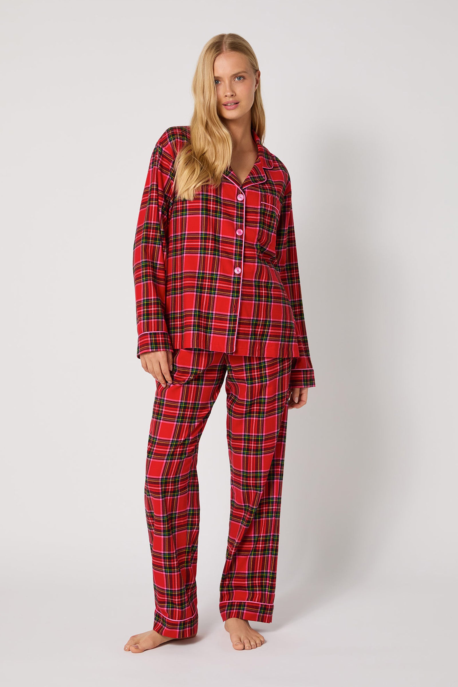 A woman with long blonde hair poses barefoot against a white background, wearing the Christmas Plaid L/S Classic Pj—a matching red plaid pajama set with long sleeves and pants.