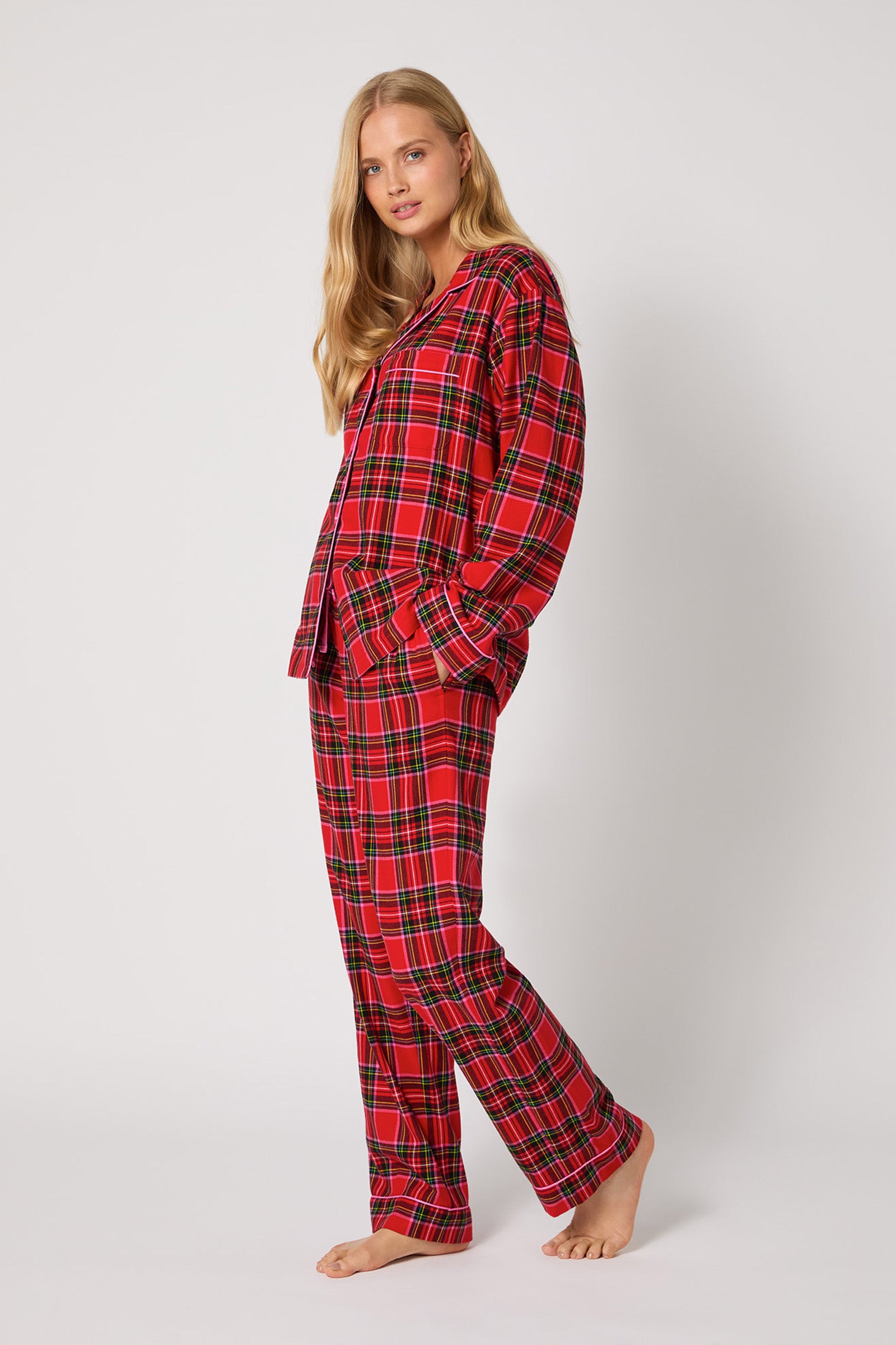 A woman with long blonde hair wears the Christmas Plaid L/S Classic PJ in red and black plaid, standing barefoot against a white background with one hand in her pocket, looking at the camera.