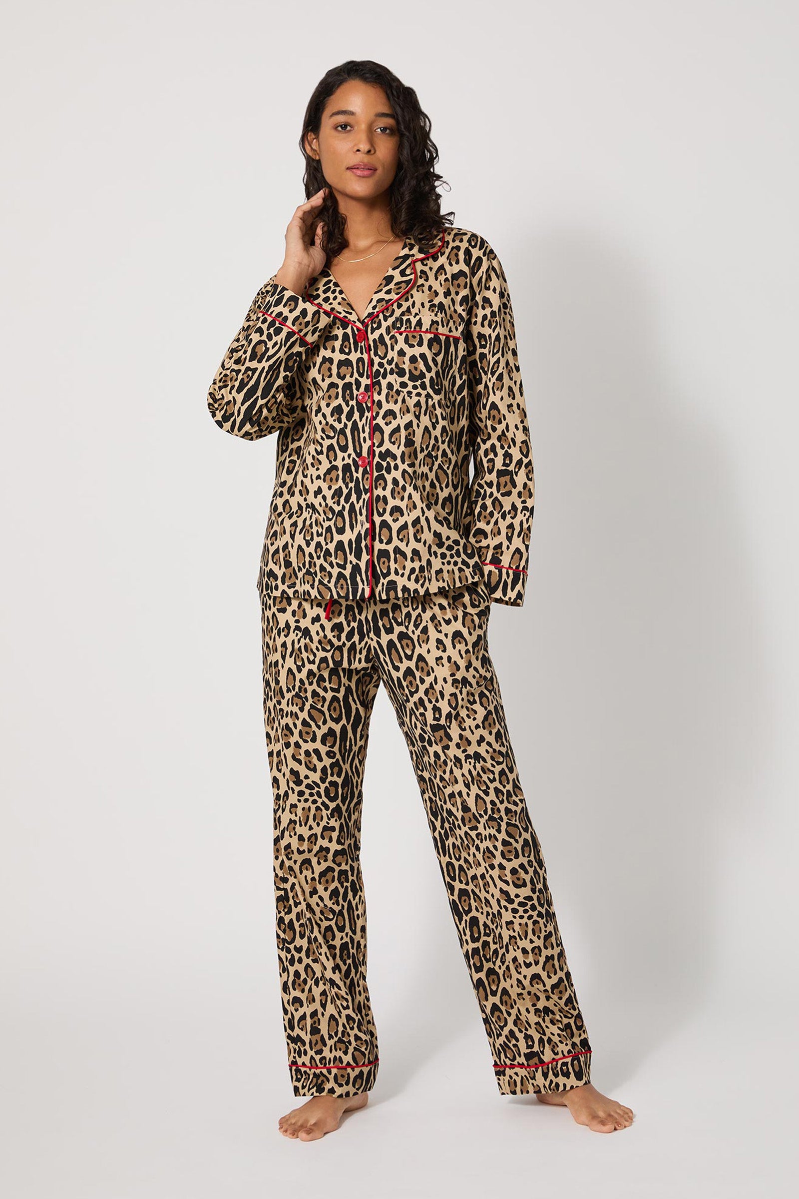 A woman stands barefoot against a white background, wearing the Leopard Stripe L/S Classic Pj—a matching long-sleeve, button-up leopard print pajama set with red accents—and touches her neck with one hand.