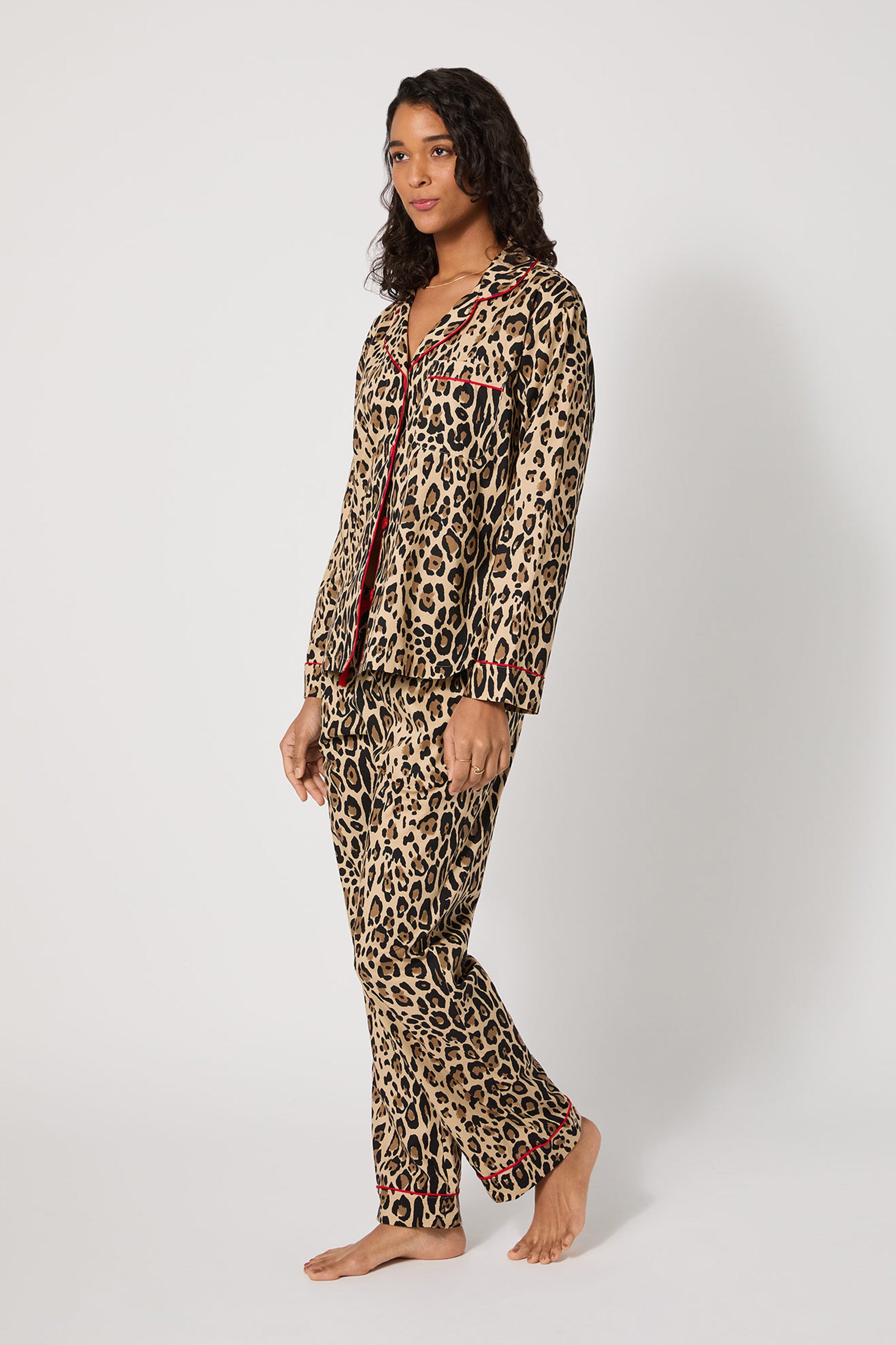 A woman stands barefoot in the Leopard Stripe L/S Classic Pj—matching leopard print pajamas with red trim, a long-sleeve button-up top, and pants—against a plain white background.