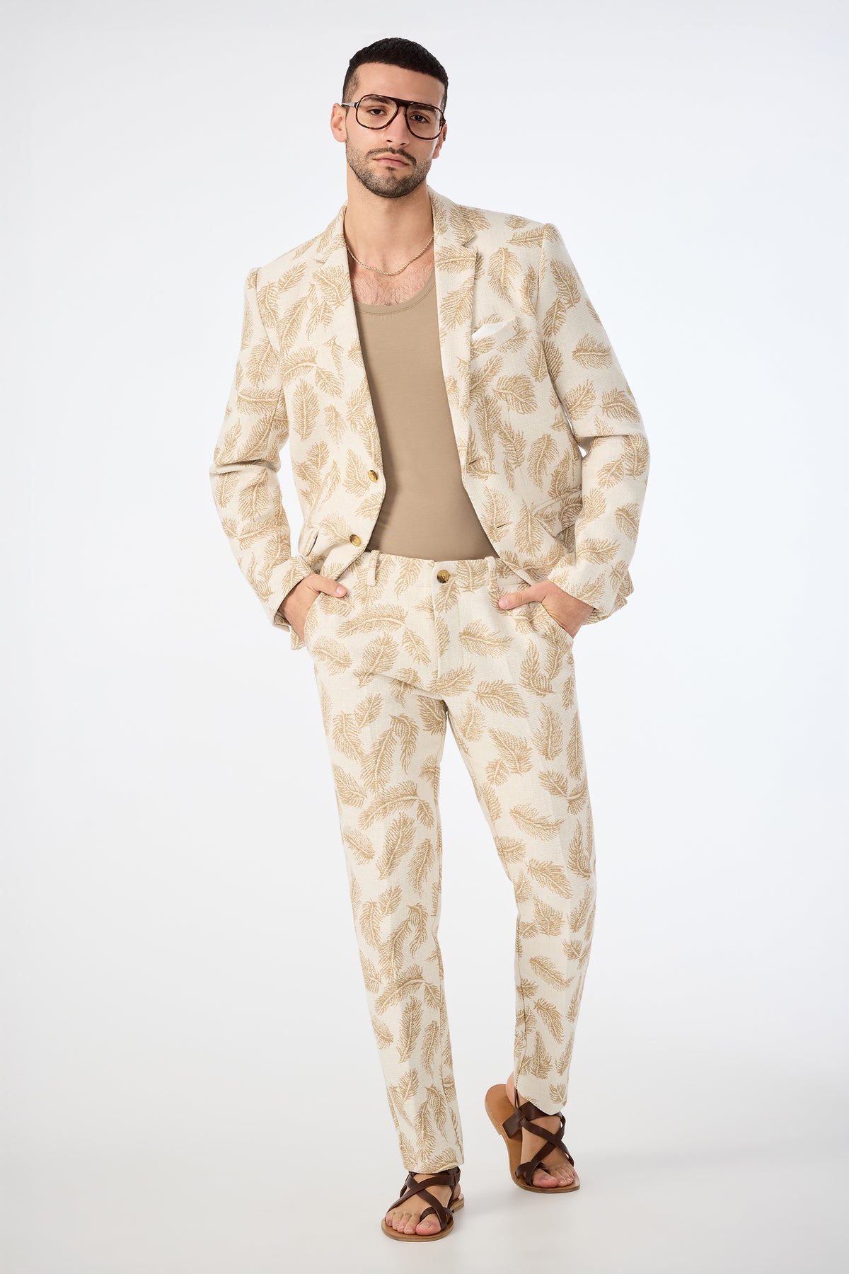 A man poses confidently against a plain white background, hands in pockets, wearing the Alex Slim Trouser in Desert Fronds Jacquard paired with a tan shirt, brown sandals, and large glasses.