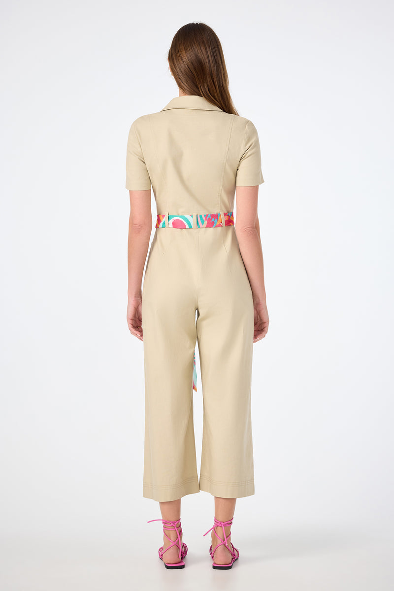 A woman faces away, wearing the JAdore V-Neck Jumpsuit in beige with short sleeves and wide legs, styled with a colorful print belt and pink strappy sandals. Her straight brown hair is down against a plain white background.