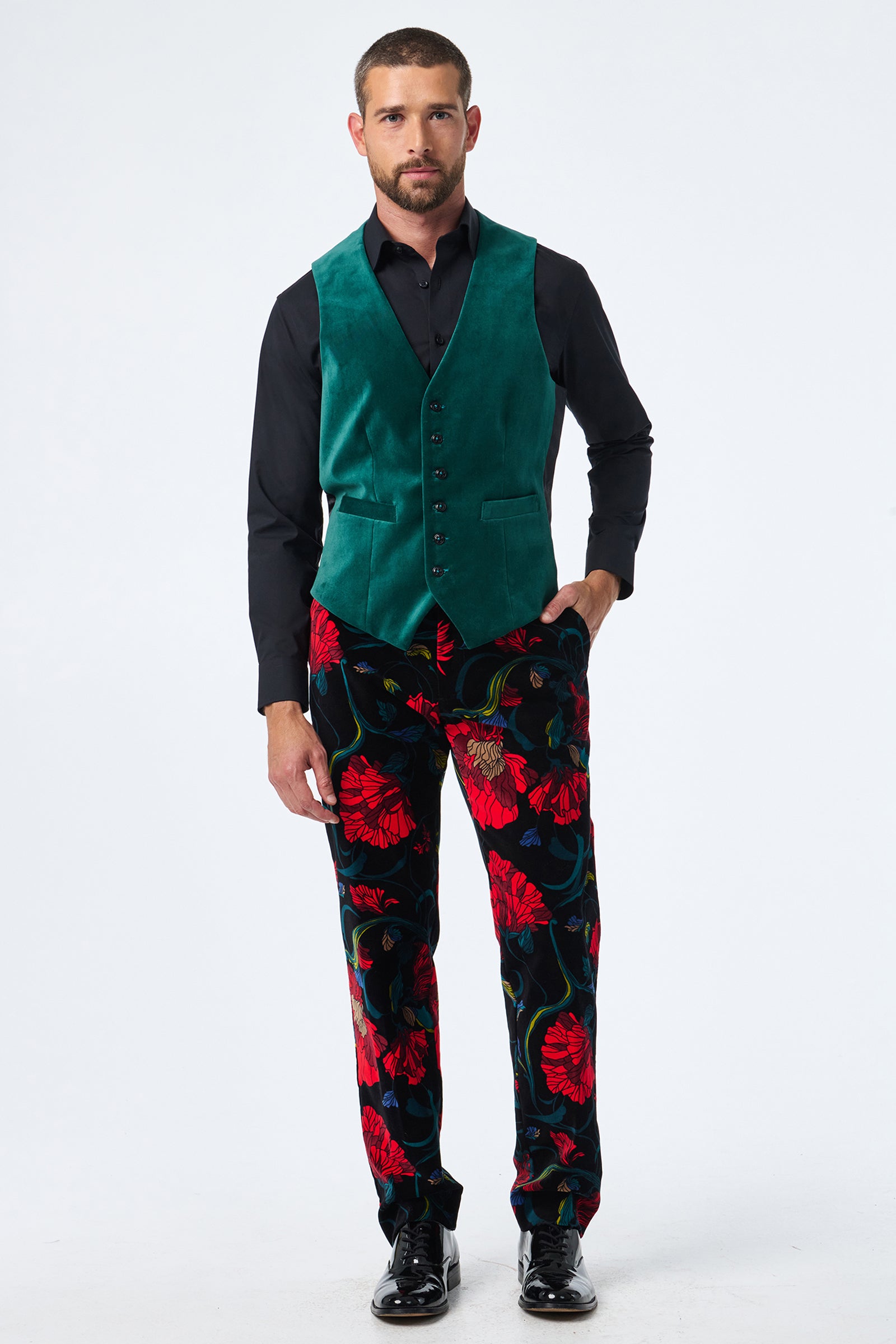 A man stands facing forward, wearing the Irving Vest in green velvet over a black shirt and bold black pants with a red floral pattern. He has short hair, a beard, and is wearing black shoes.