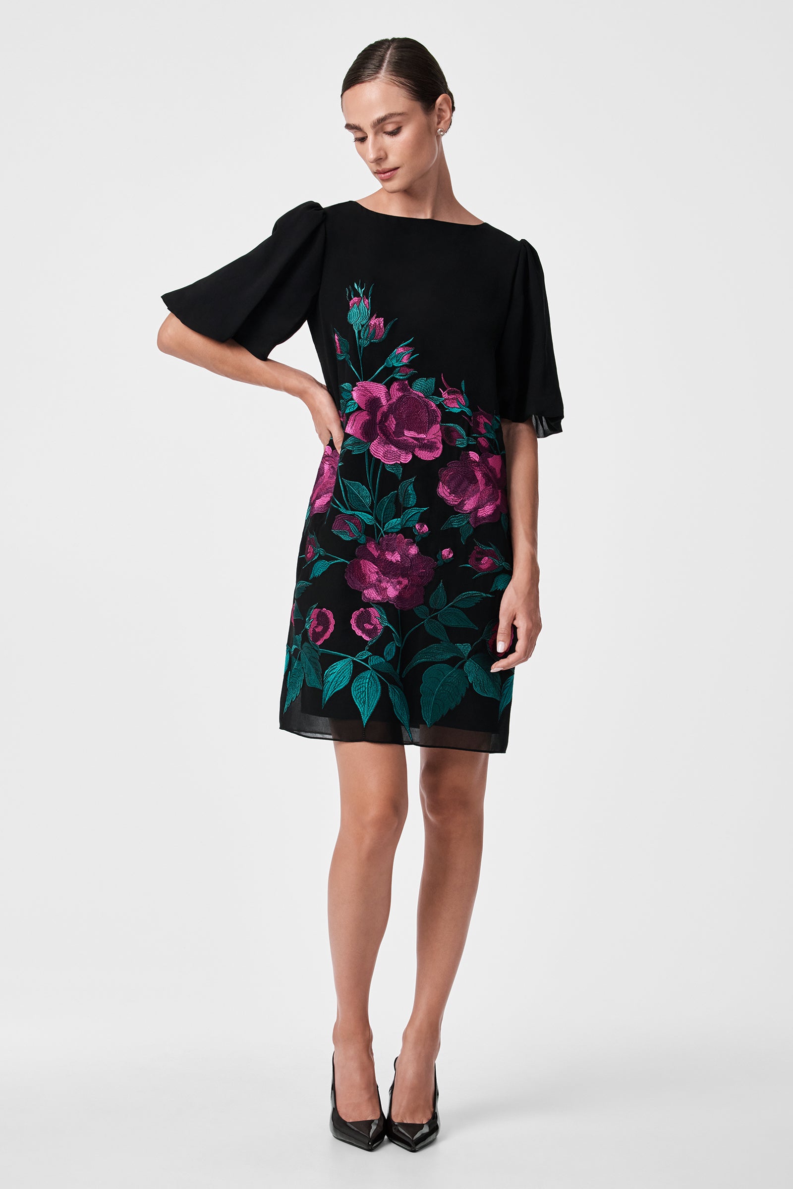 A woman stands in black heels against a white background, wearing the Luv Dress—a black dress with flutter sleeves and a bold pink and green floral print on the front.