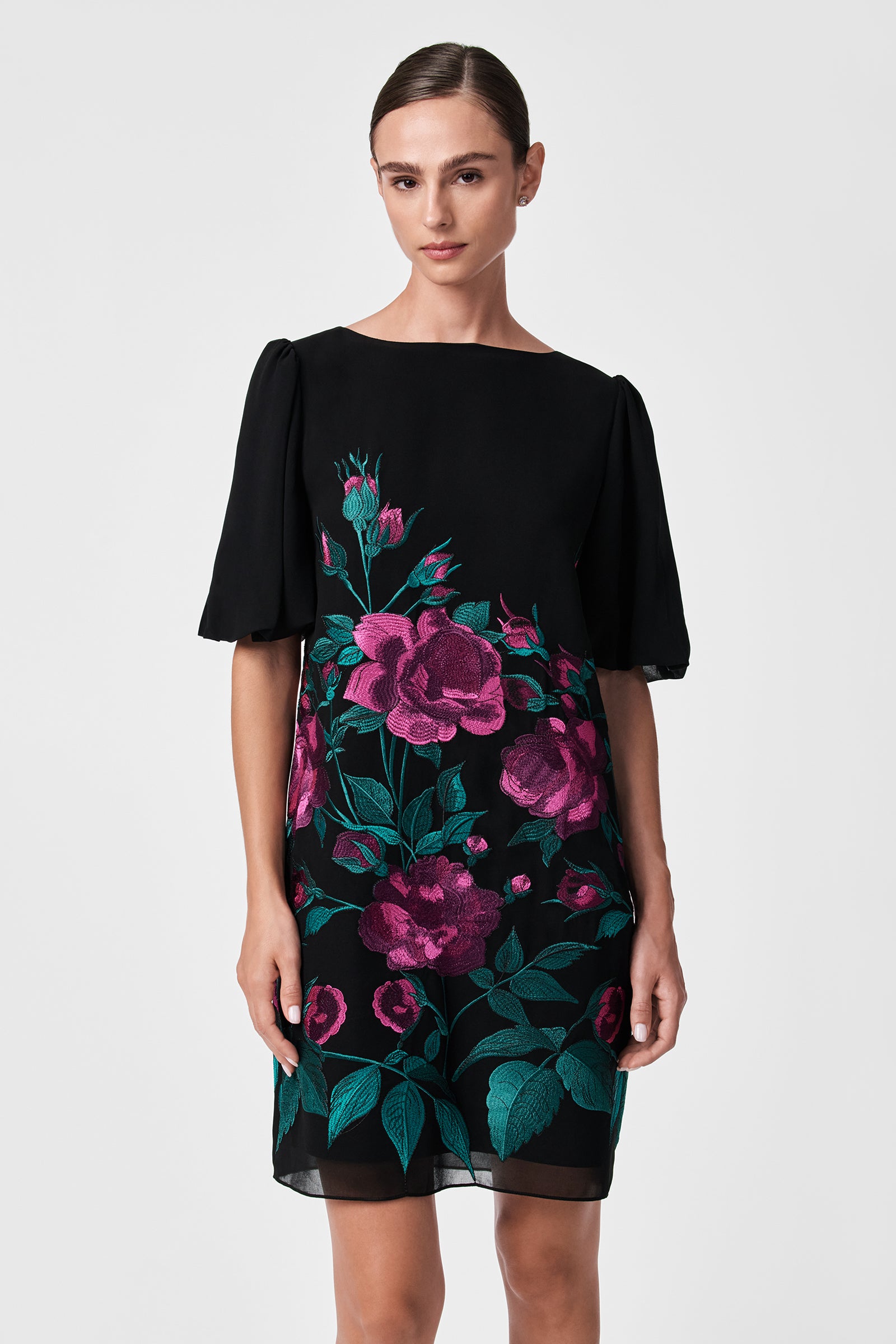 A woman models the Luv Dress, a black dress with short, loose sleeves and vibrant pink and green floral embroidery on the front, standing against a plain light gray background.