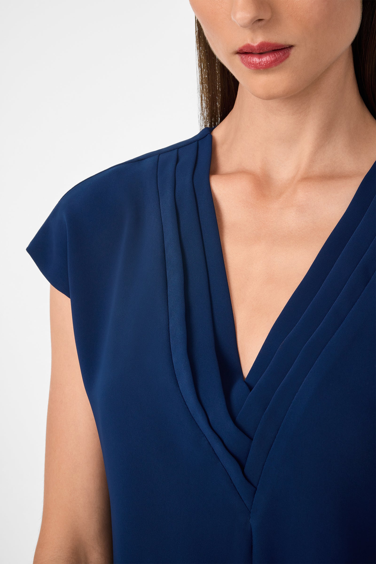 A woman models the Zula Top, a deep blue blouse with a structured V-neckline and short sleeves; only her lower face, neck, and upper torso appear against a plain background.