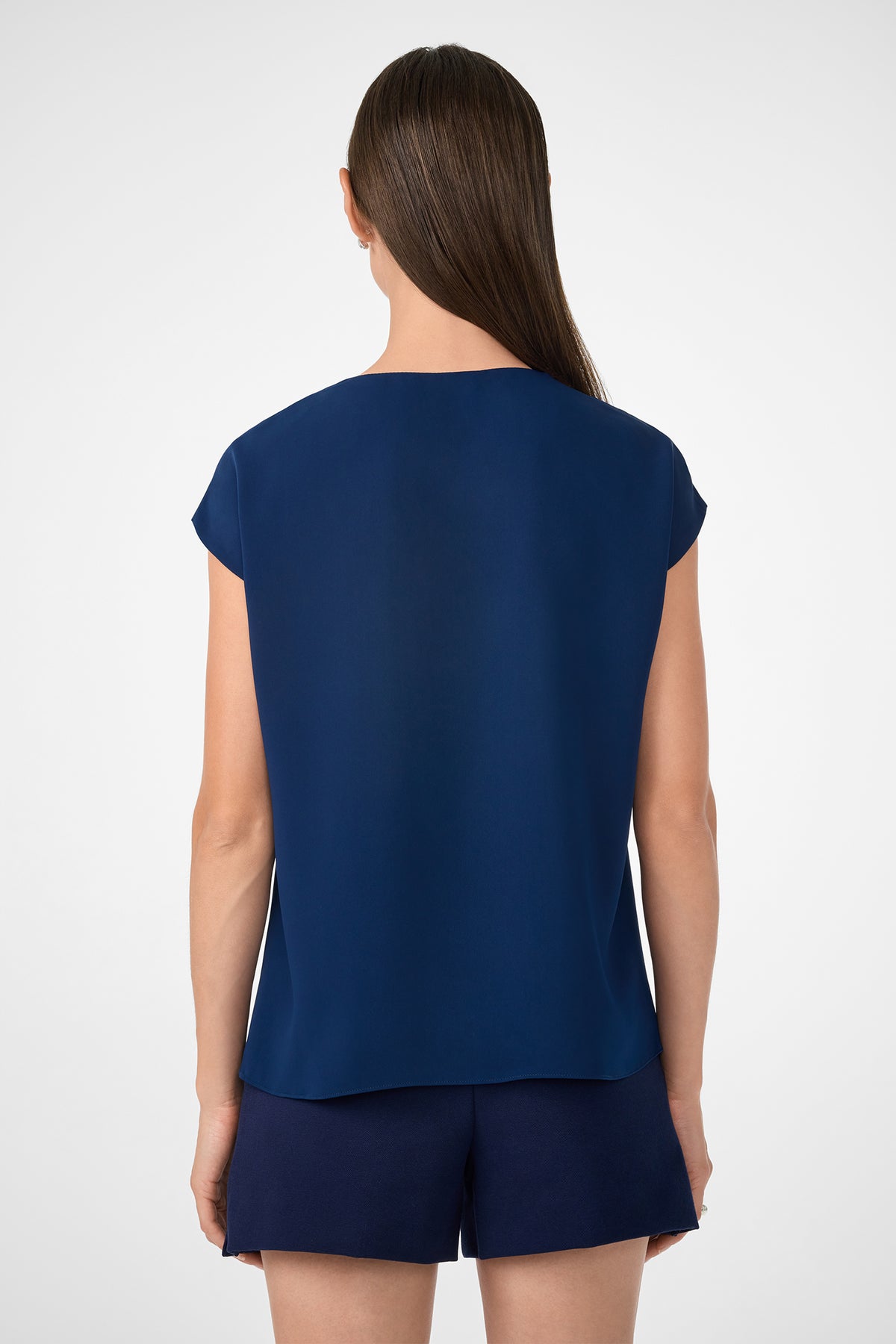 A woman with straight brown hair, facing away, wears the Zula Top—a loose-fitting, short-sleeve dark blue top—paired with matching shorts against a plain light background.