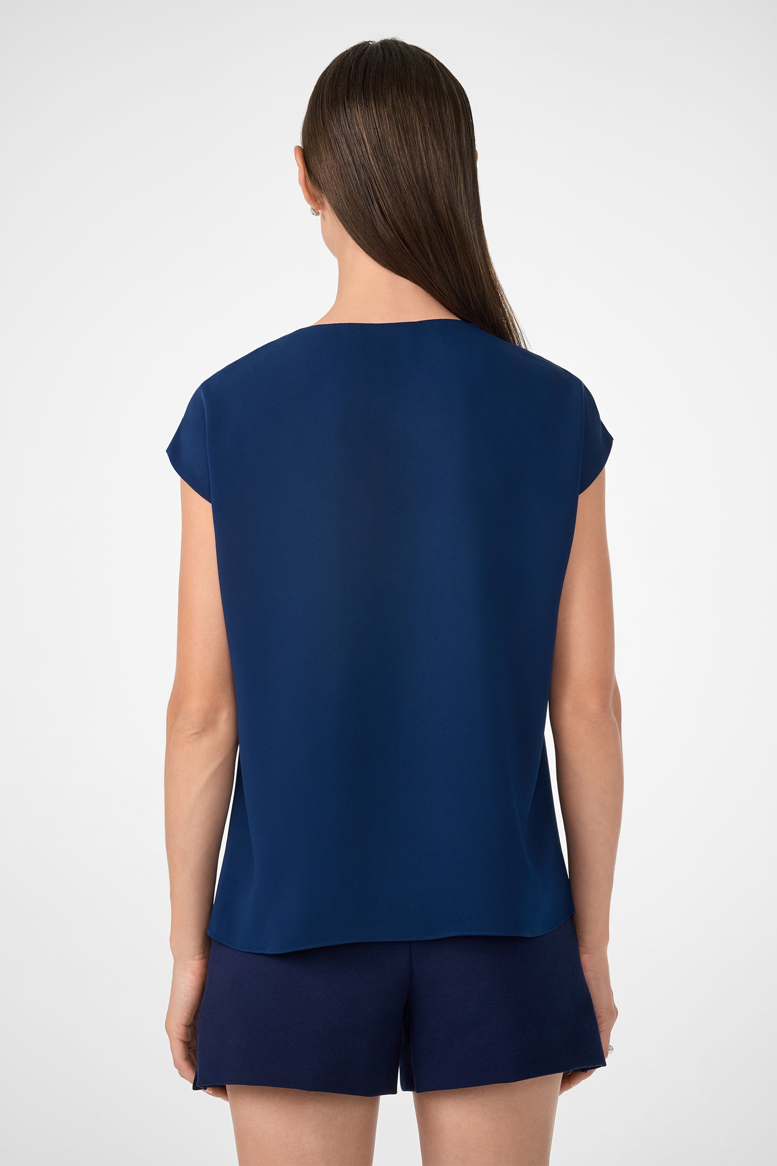 A woman with straight brown hair, facing away, wears the Zula Top—a loose-fitting, short-sleeve dark blue top—paired with matching shorts against a plain light background.