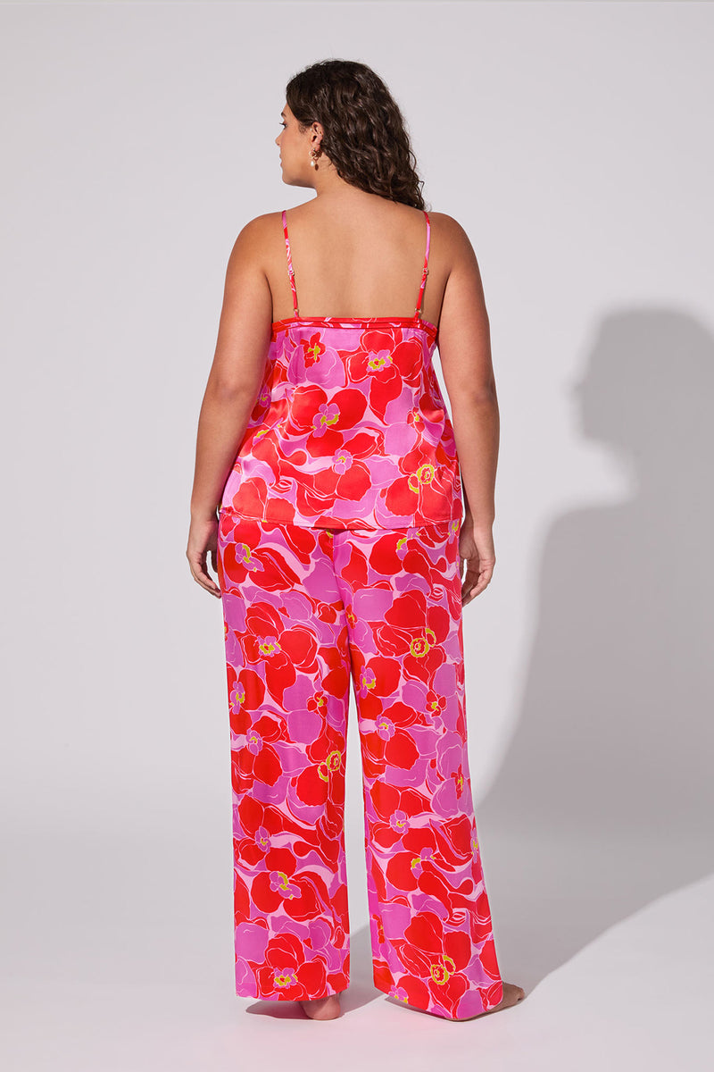 A woman stands barefoot, facing away, wearing the Silk Cami PJ Set - Winter Orchids: a matching floral set with loose-fitting pants and a spaghetti strap top. The background is plain light gray.