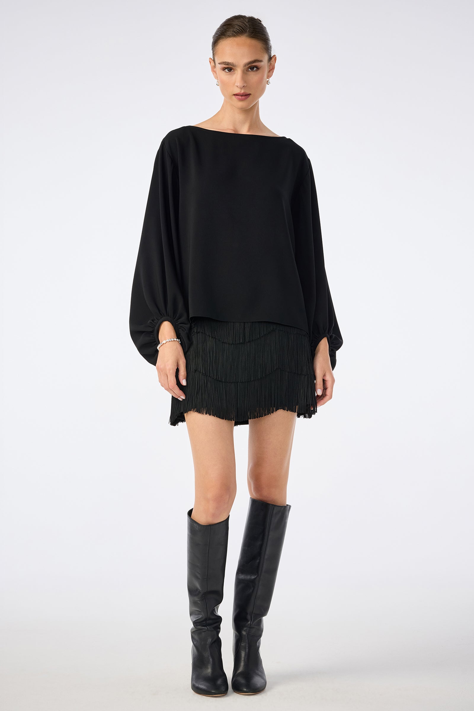 A woman faces forward, wearing the URSA 2 TOP— a loose black long-sleeve top—paired with a black fringed polyester mini skirt and knee-high black boots, set against a plain white background.