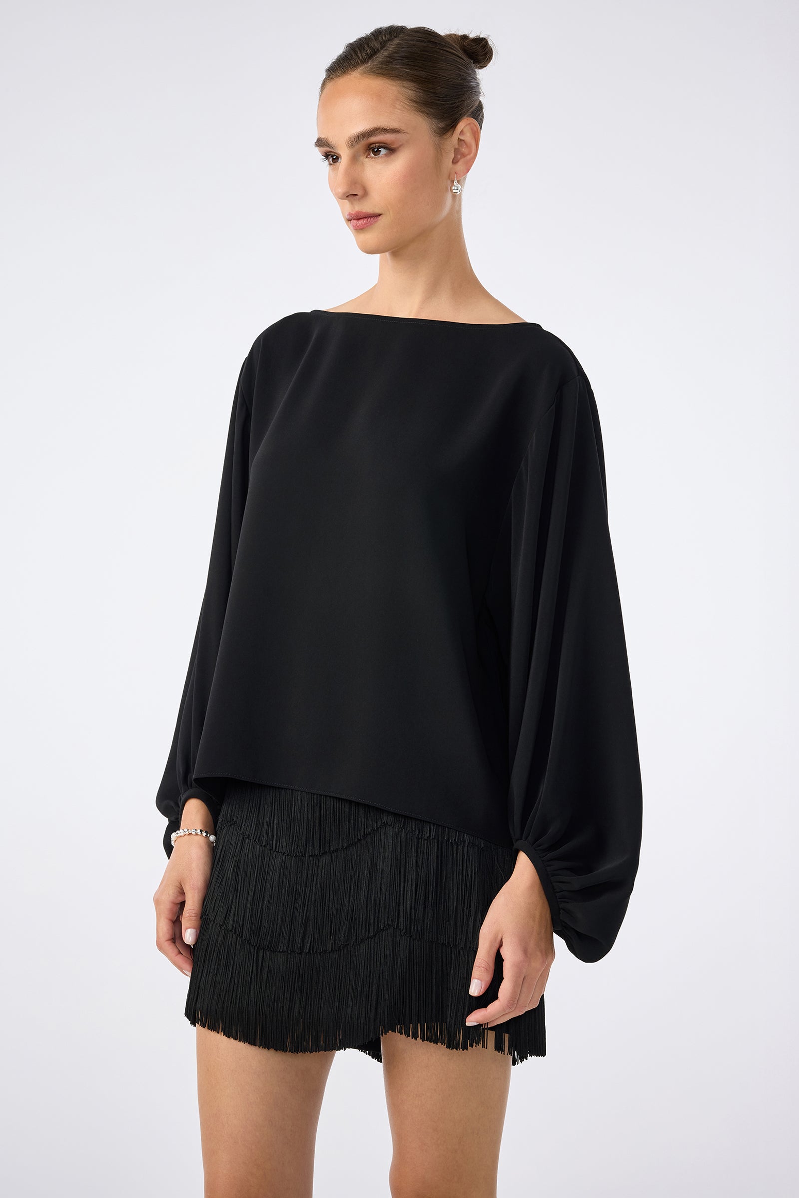A woman wearing the URSA 2 TOP, a black long-sleeved top with voluminous sleeves, and a black fringed 100% polyester mini skirt stands against a plain light background, gazing slightly to the side.