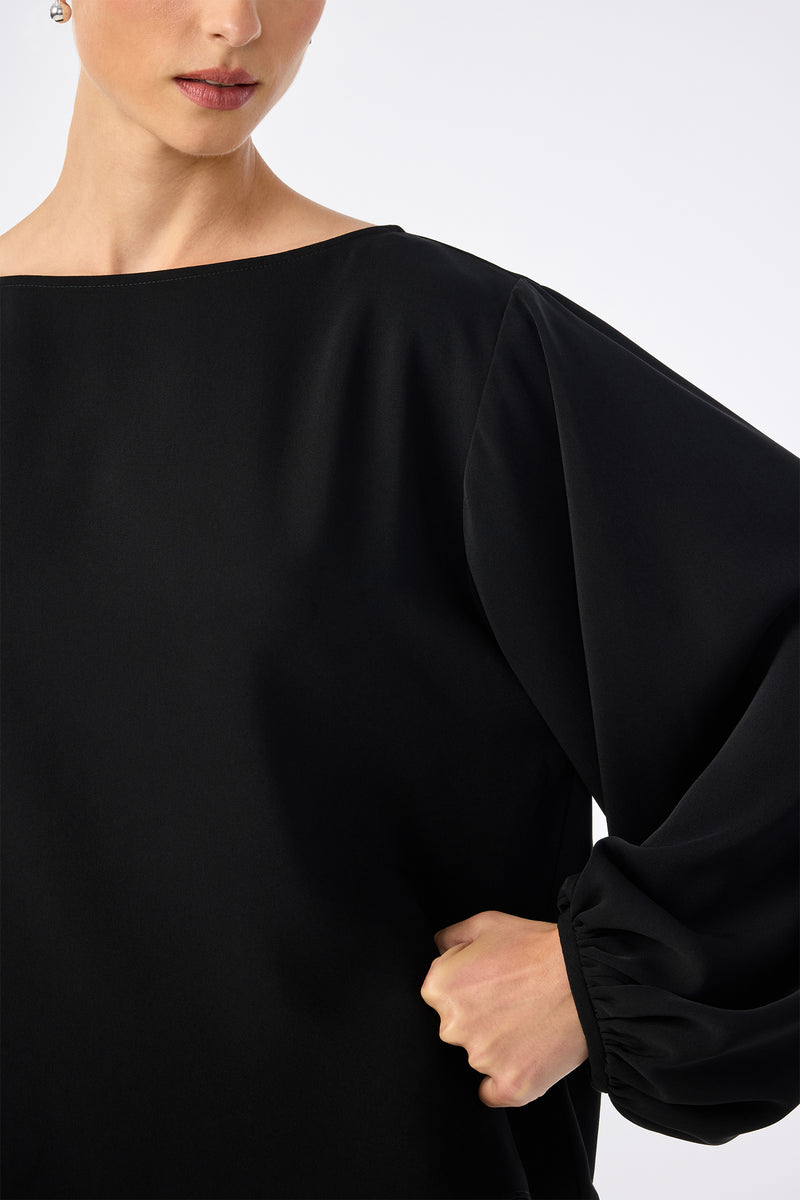 A woman models the URSA 2 TOP, a loose-fitting black long-sleeve blouse with puffed sleeves made from 100% polyester, shown from shoulders to waist, one hand on her hip, against a plain light background.