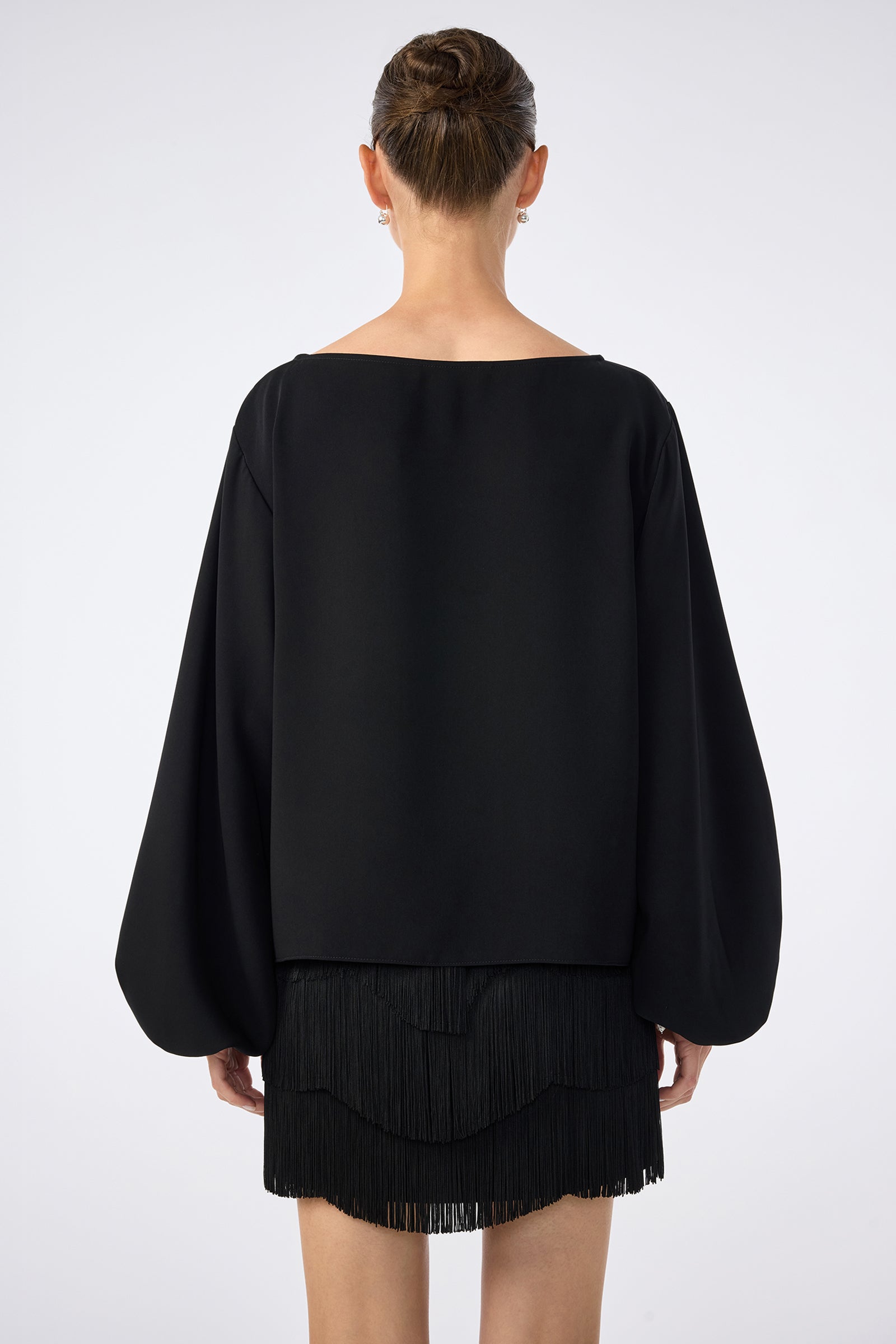 A woman with her hair in a neat bun faces away, wearing the URSA 2 TOP—a loose-fitting black top with wide sleeves made from 100% polyester—paired with a layered fringe skirt, set against a plain white background.