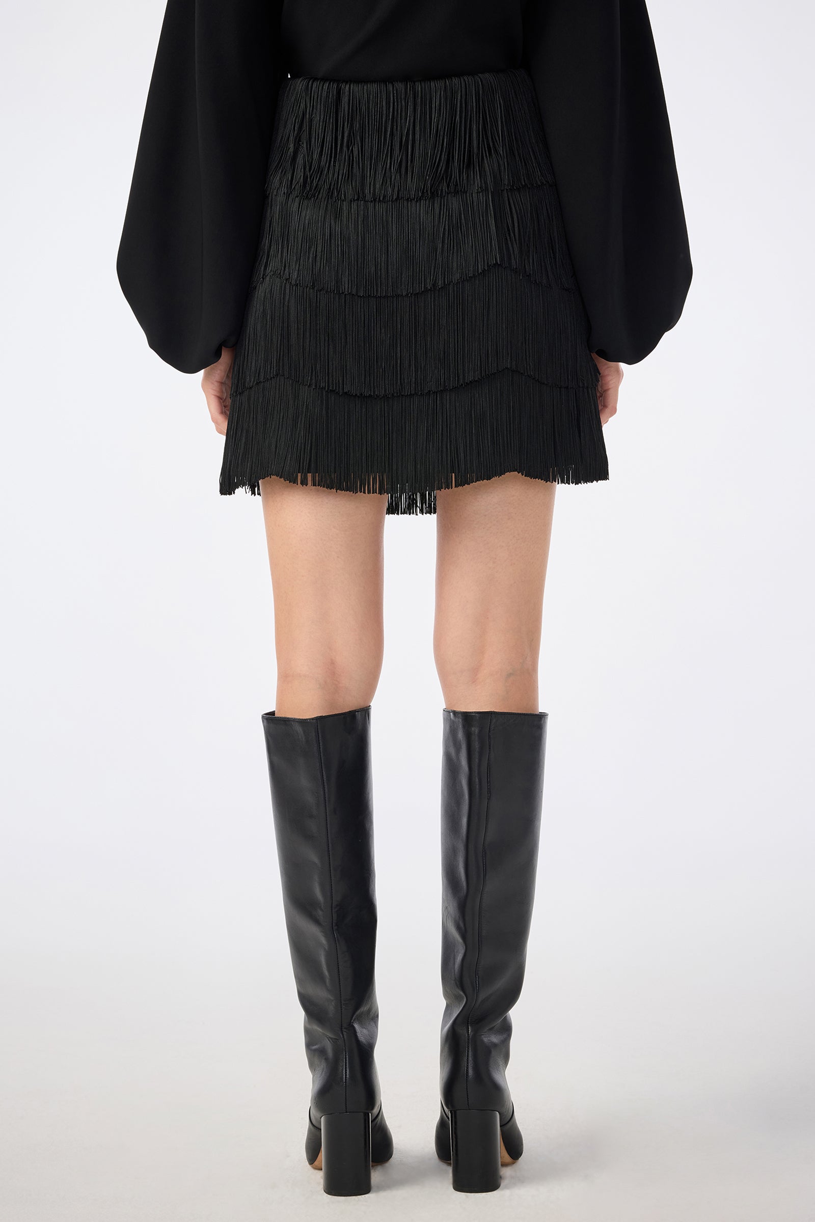 A person stands facing away against a white background, wearing the Chocolat Skirt—a black fringed mini skirt—paired with a black long-sleeve top with wide sleeves and knee-high black block-heeled boots.