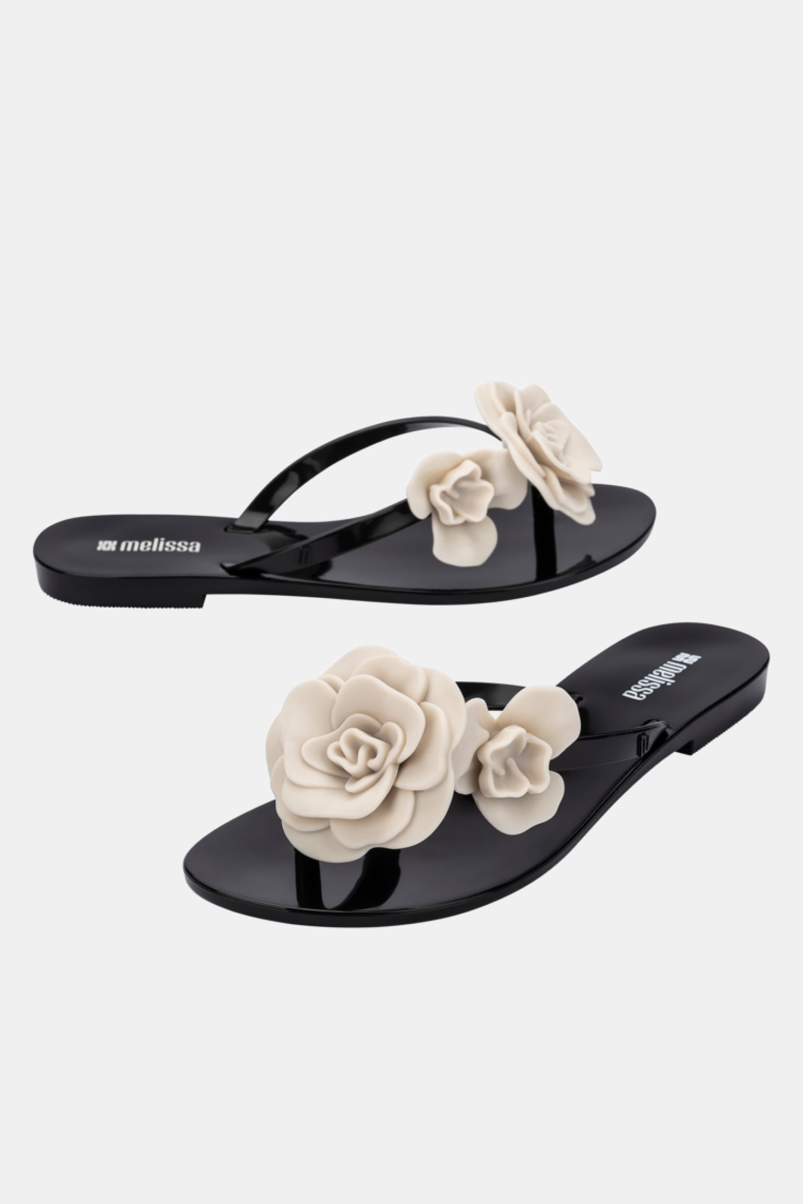 The Melissa Harmonic Petals Sandal is a pair of glossy black flip-flops featuring large, beige 3D flower decorations on the straps, displayed against a white background.