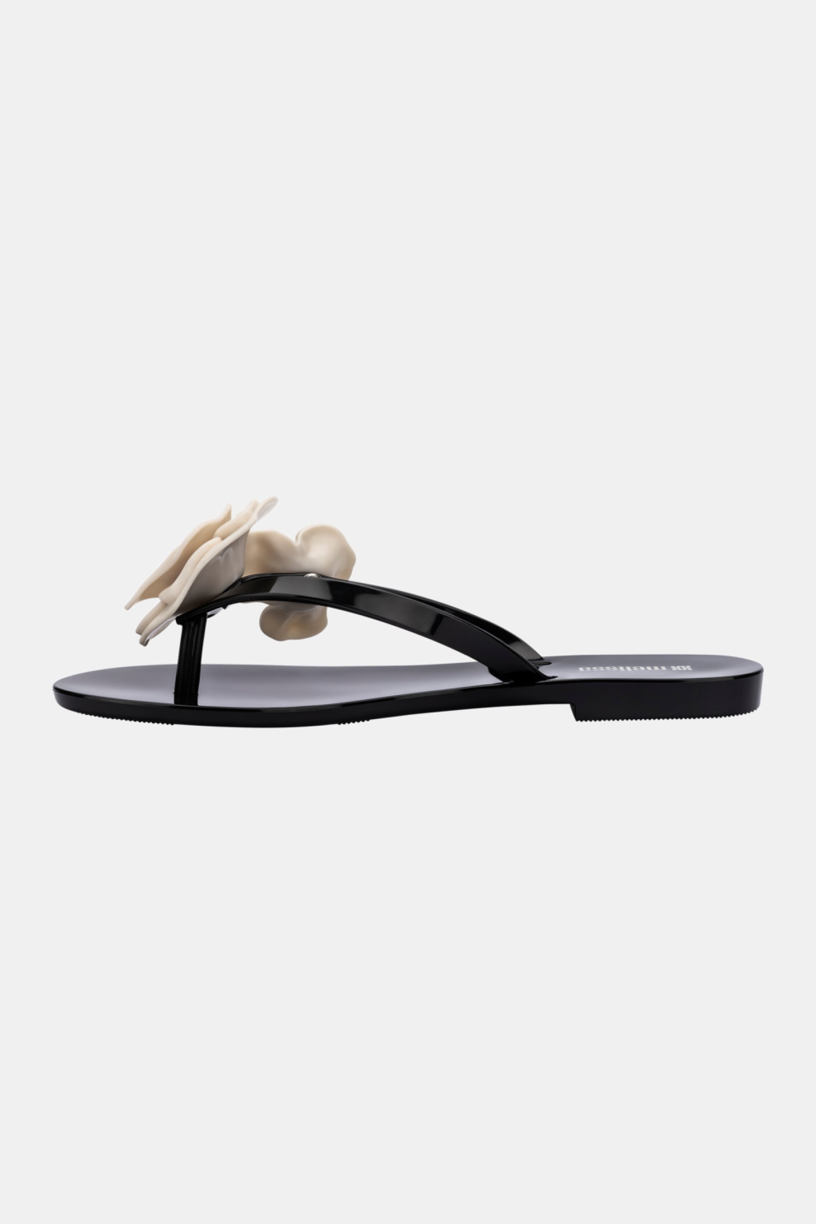 The Melissa Harmonic Petals Sandal is a black, glossy flat sandal with a minimalist thong design, featuring a large cream flower on the strap.