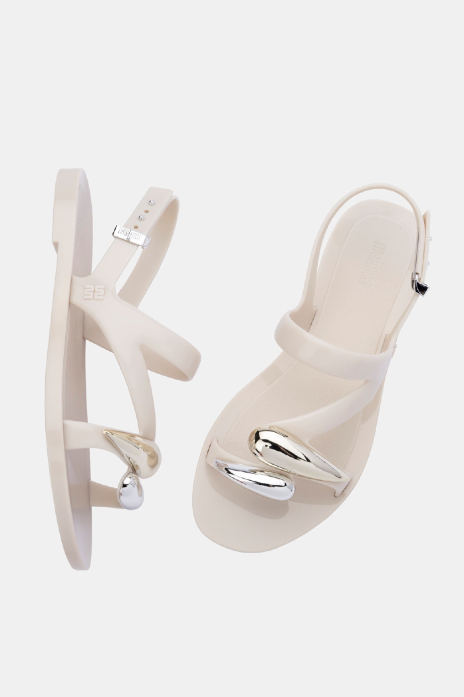 The Melissa Iris Sandal features beige heels with clear straps and shiny silver sculptural accents on the toes, shown against a plain white background.