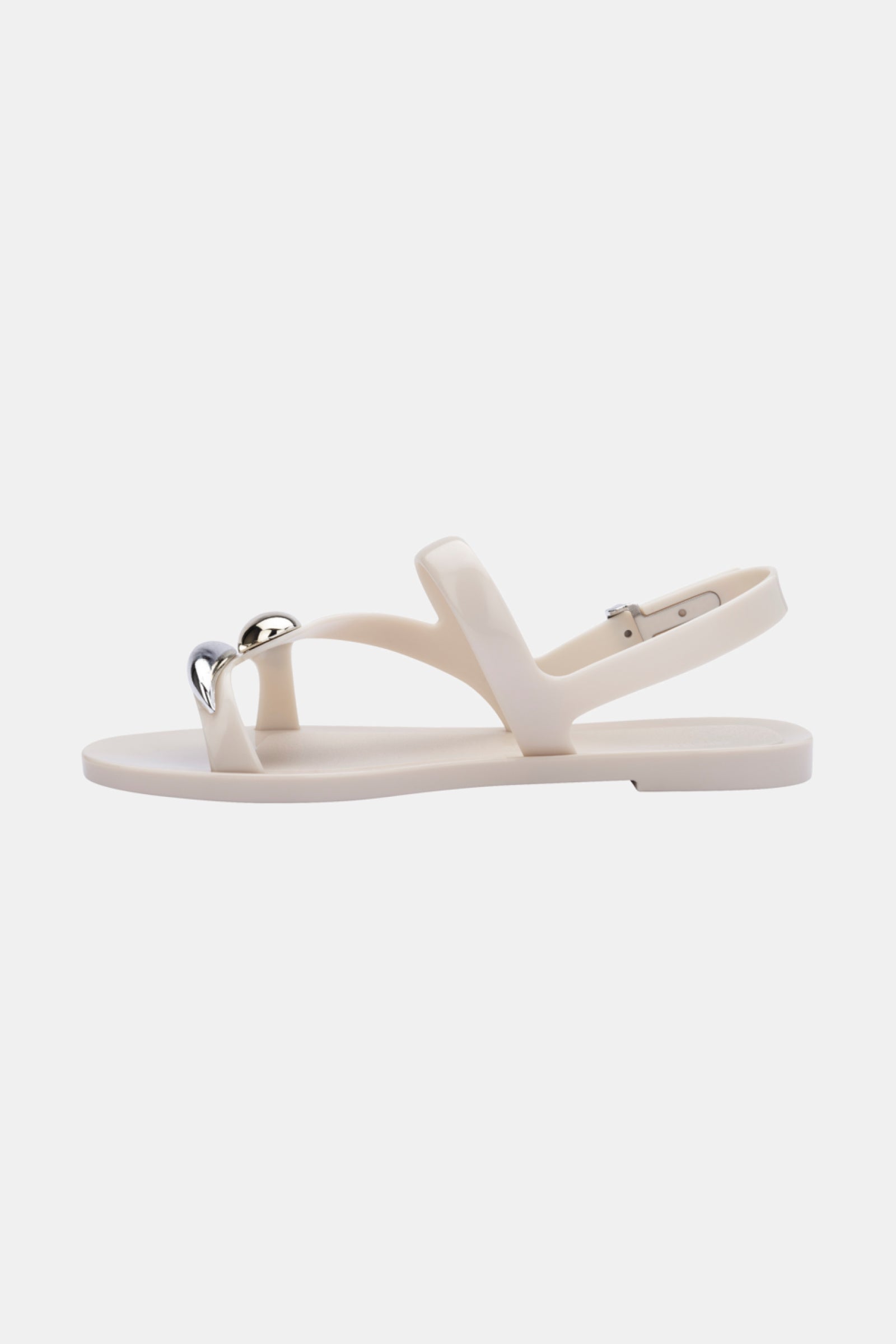 The Melissa Iris Sandal is a minimalist white sandal with a flat sole, toe strap featuring a silver heart-shaped detail, and an adjustable ankle strap, shown on a plain white background.