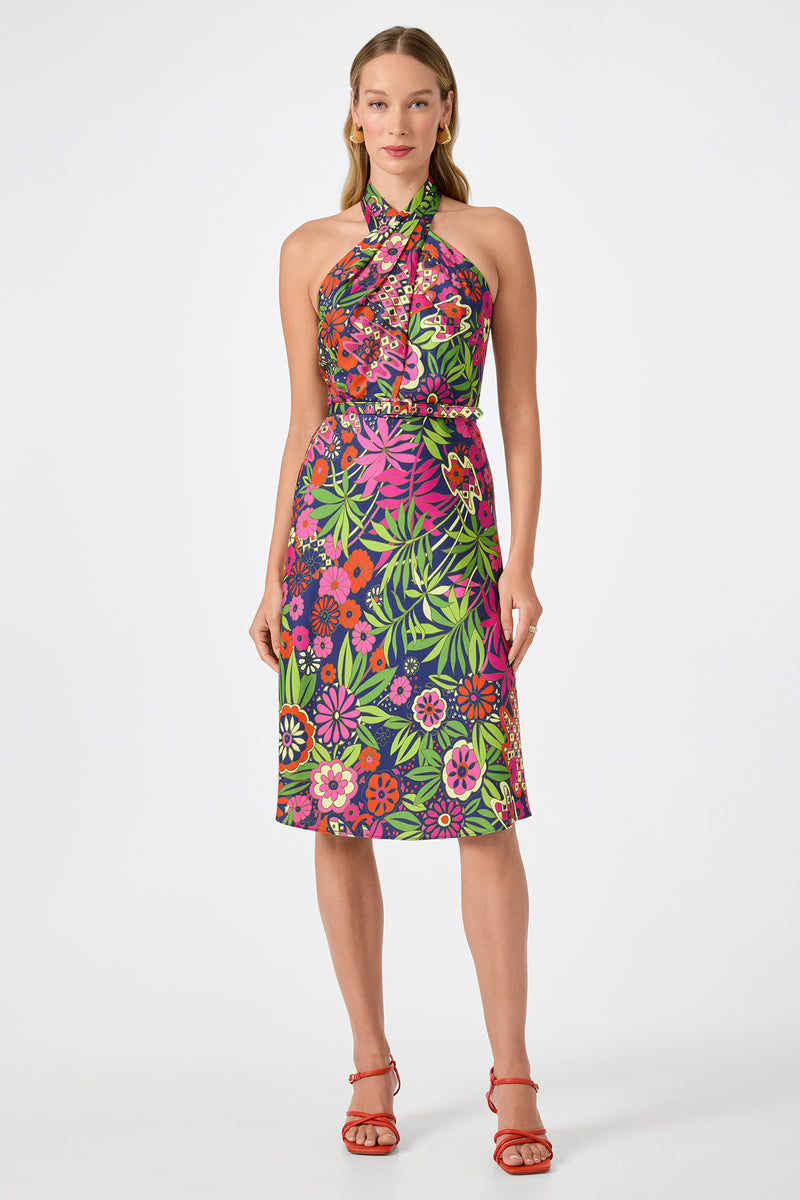 A woman models the Muri Cross-Halter Midi Dress in Fumi Floral, featuring a vibrant pink, green, and purple pattern. She wears red strappy sandals and stands against a plain white background.