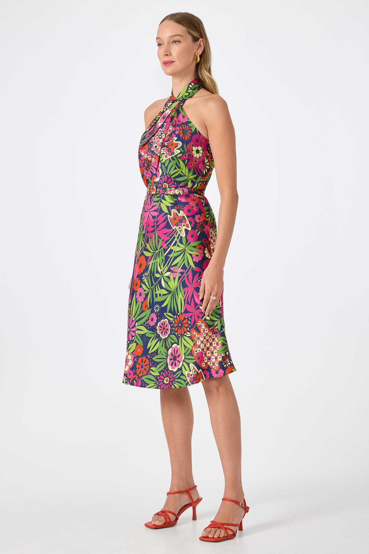 A woman poses against a plain white background in the Muri Cross-Halter Midi Dress - Fumi Floral, a sleeveless, knee-length dress with bright pink, green, and purple patterns. She completes the look with red strappy heels and long, straight hair.