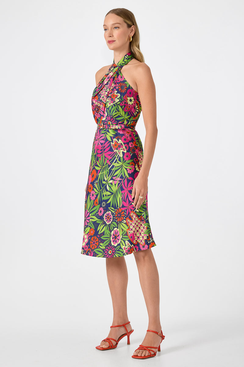 A woman poses against a plain white background in the Muri Cross-Halter Midi Dress - Fumi Floral, a sleeveless, knee-length dress with bright pink, green, and purple patterns. She completes the look with red strappy heels and long, straight hair.