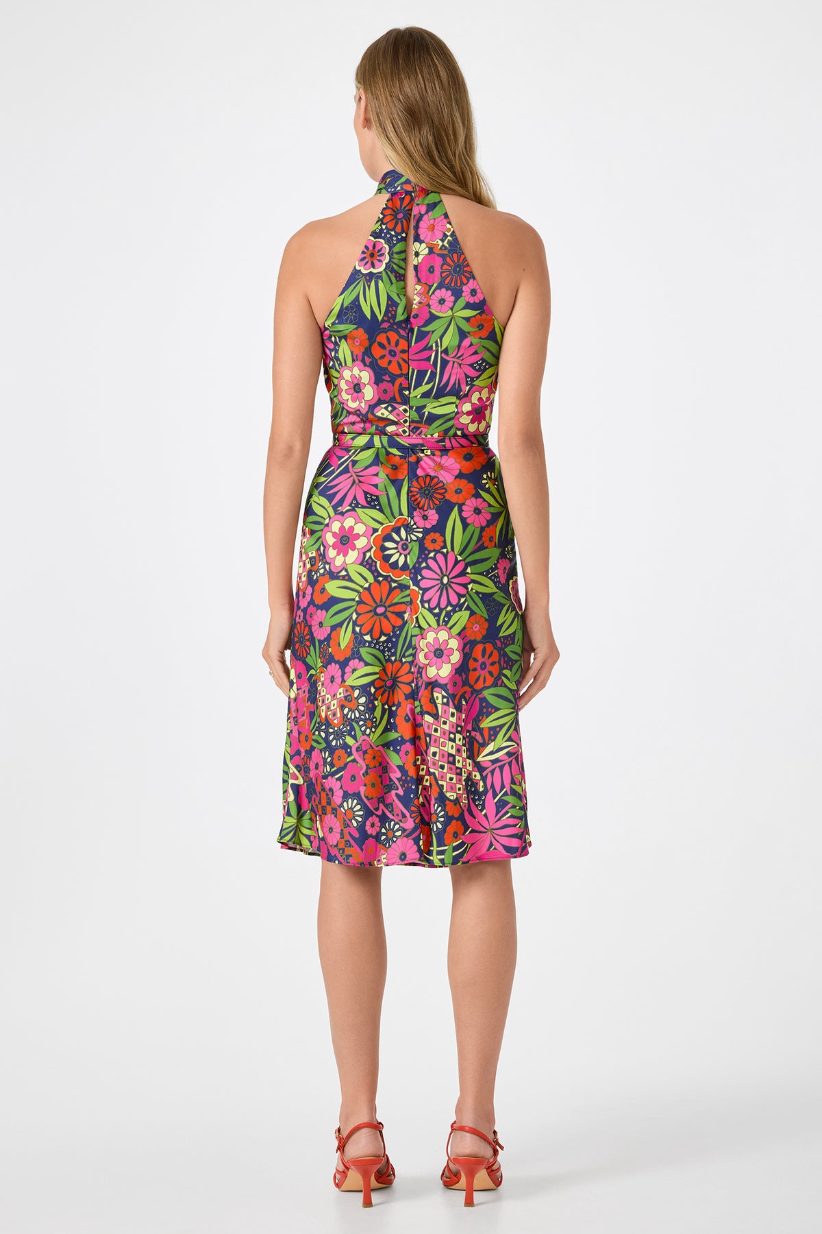 A woman stands facing away, wearing the Muri Cross-Halter Midi Dress - Fumi Floral, a colorful floral halter-neck dress thats fitted at the waist and knee-length, paired with red heeled sandals. The background is plain white.