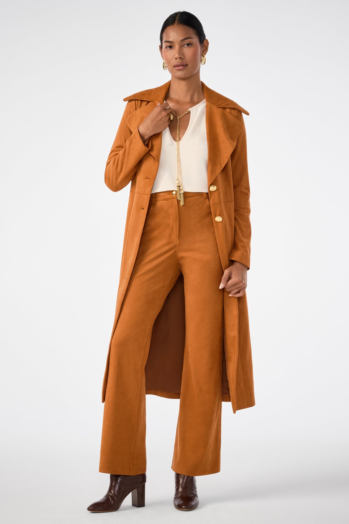 A woman stands confidently against a white background wearing the Apple Bottom Pant in rust with a matching long coat, cream blouse, brown heeled boots, gold jewelry, and hoop earrings.