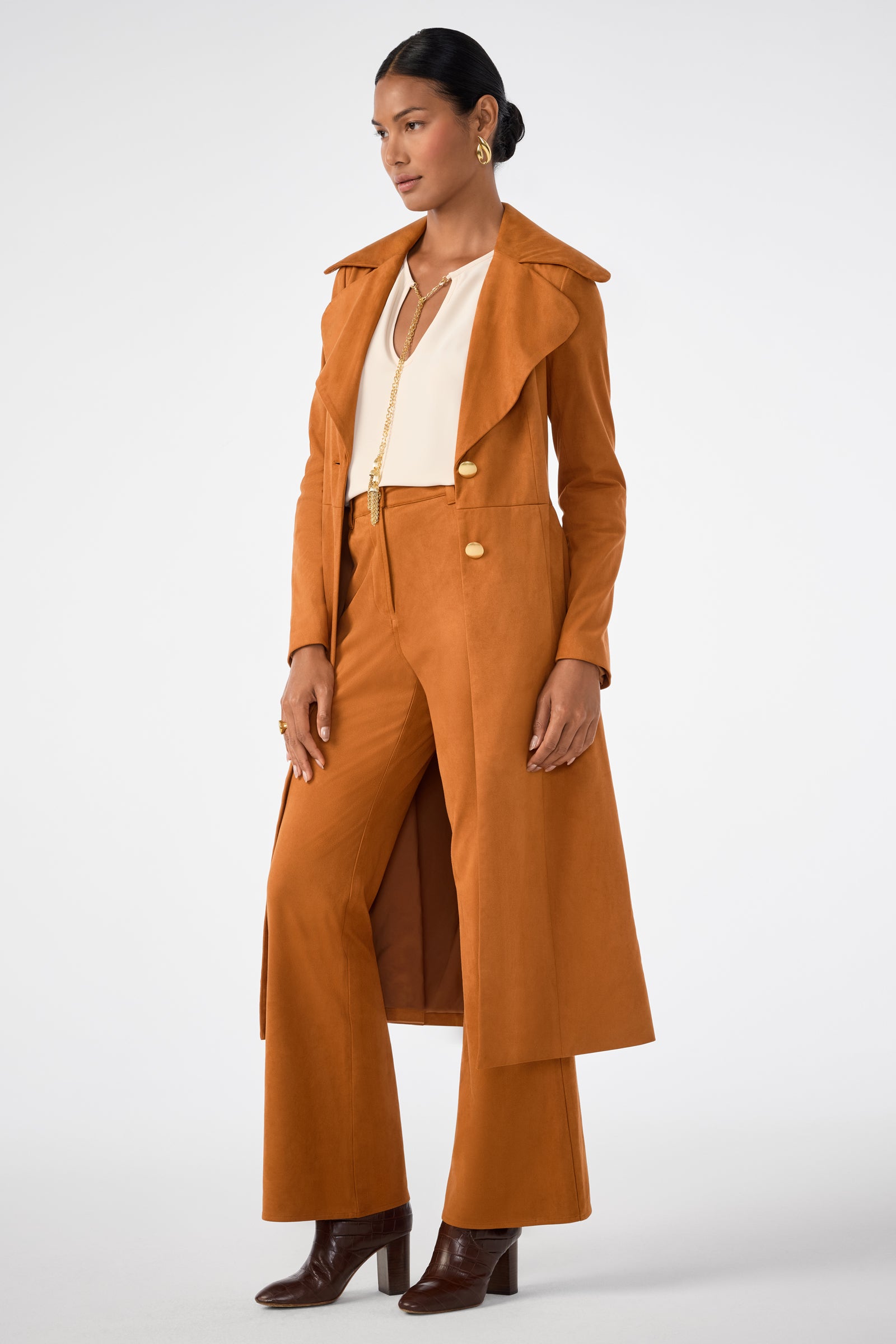 A woman stands against a plain white background wearing the Starflower Coat in rust, paired with matching wide-leg pants, a cream blouse, brown heeled boots, and gold hoop earrings.