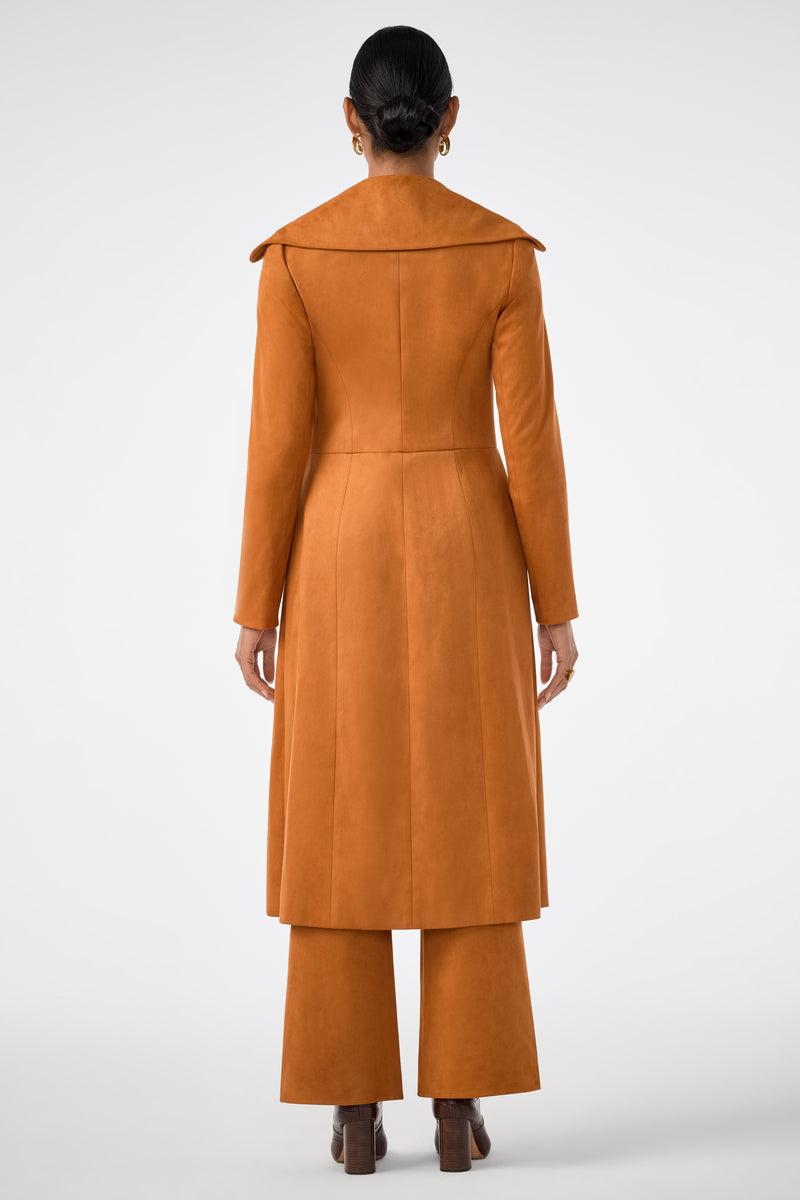 A person stands facing away, wearing the Starflower Coat—a long, burnt orange piece with a wide collar—paired with matching wide-leg pants and heeled shoes against a plain light background.