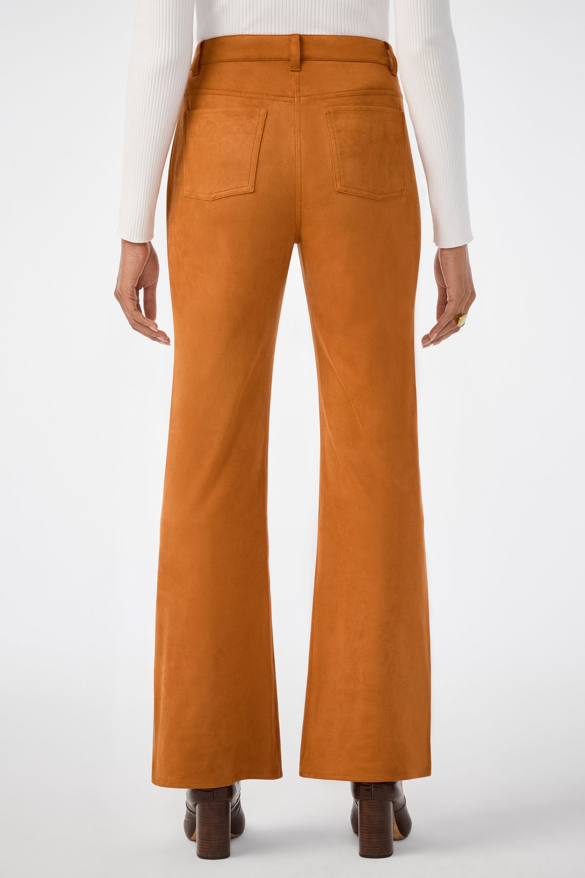 A person stands facing away from the camera wearing burnt orange Apple Bottom Pants, a white ribbed long-sleeve top, brown heeled shoes, and a ring on their right hand.