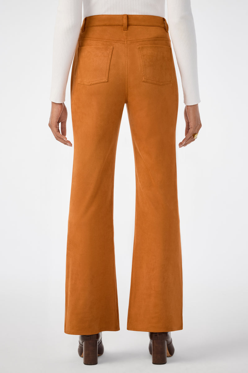 A person stands facing away from the camera wearing burnt orange Apple Bottom Pants, a white ribbed long-sleeve top, brown heeled shoes, and a ring on their right hand.