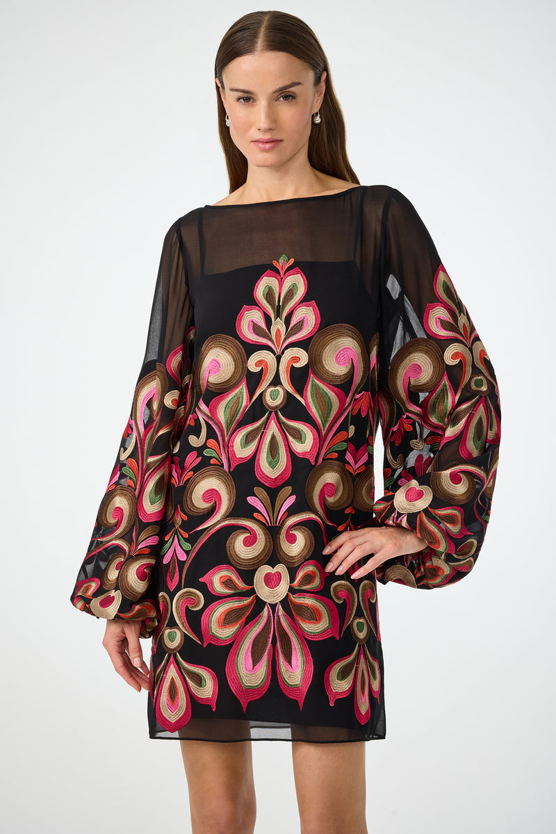 A woman stands with one hand on her hip in the Salim Embroidered Mini Dress—a black, sheer long-sleeve dress featuring bold floral and paisley embroidery in pink, green, gold, and red—against a plain background.