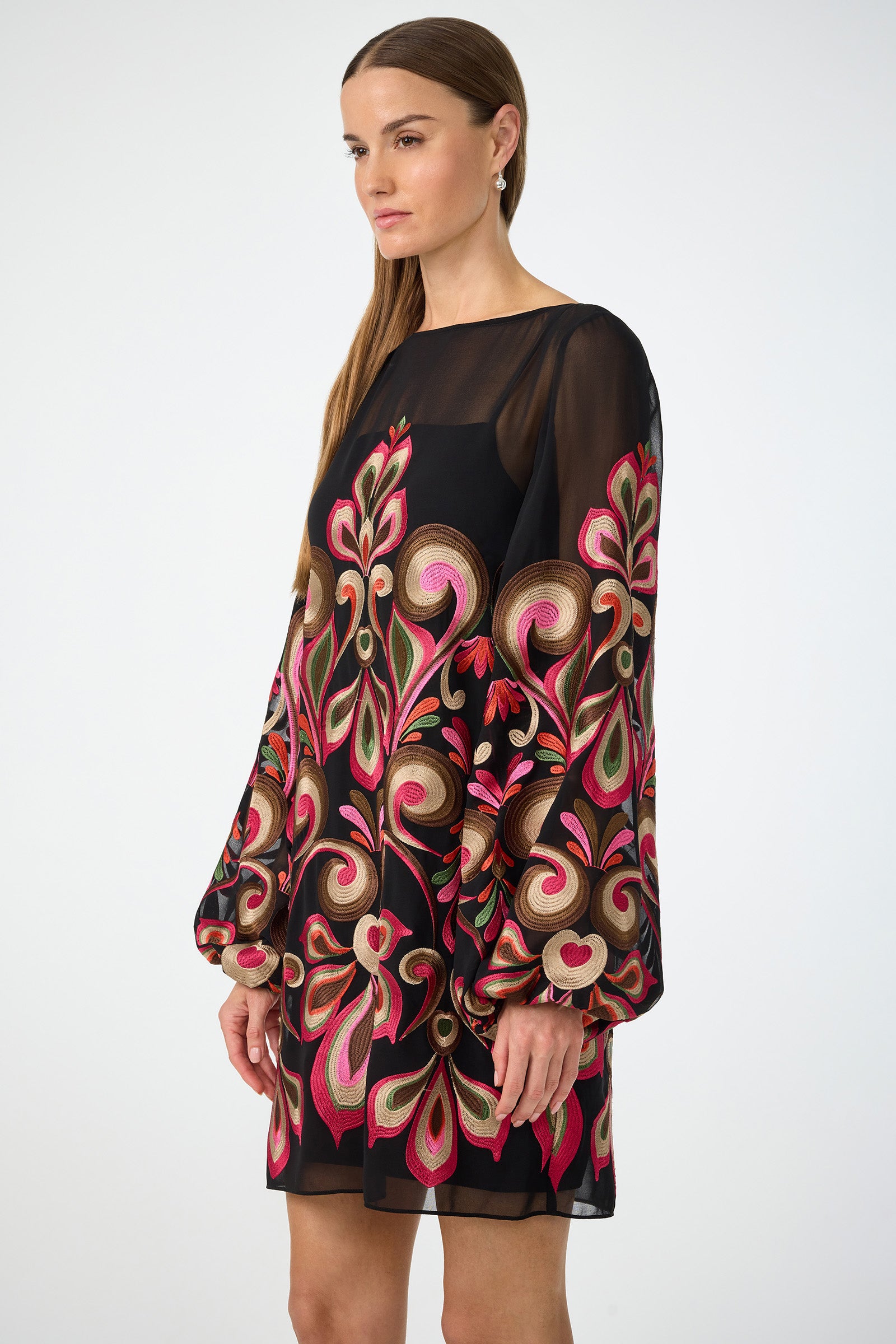A woman with straight brown hair and a neutral expression models the Salim Embroidered Mini Dress, featuring sheer black fabric, bold colorful floral embroidery, and long billowy sleeves, against a plain white background.