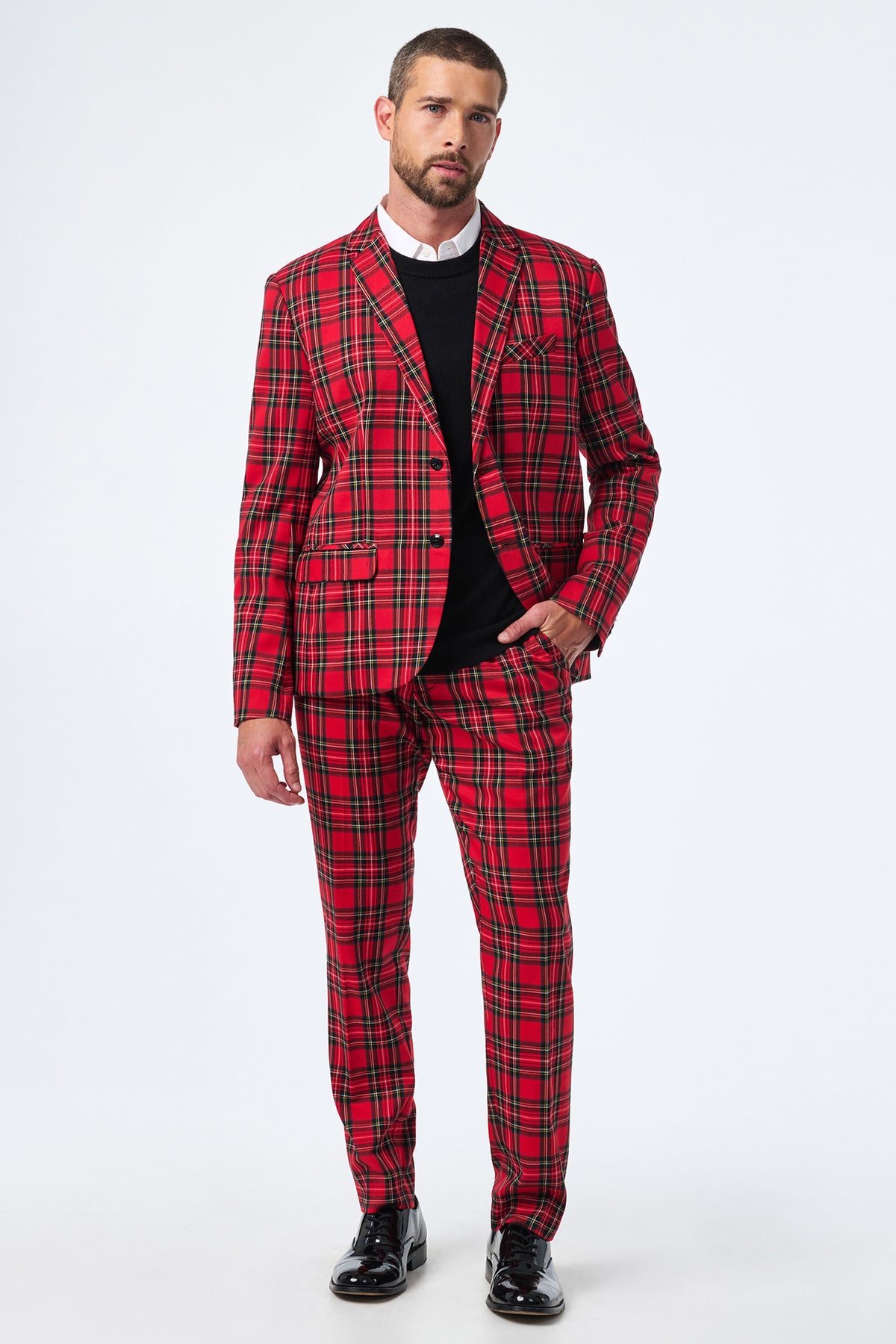 A man stands against a plain white background wearing the Thurston Tartan Blazer—a red and black plaid suit jacket—paired with a white shirt, black sweater, and black dress shoes.