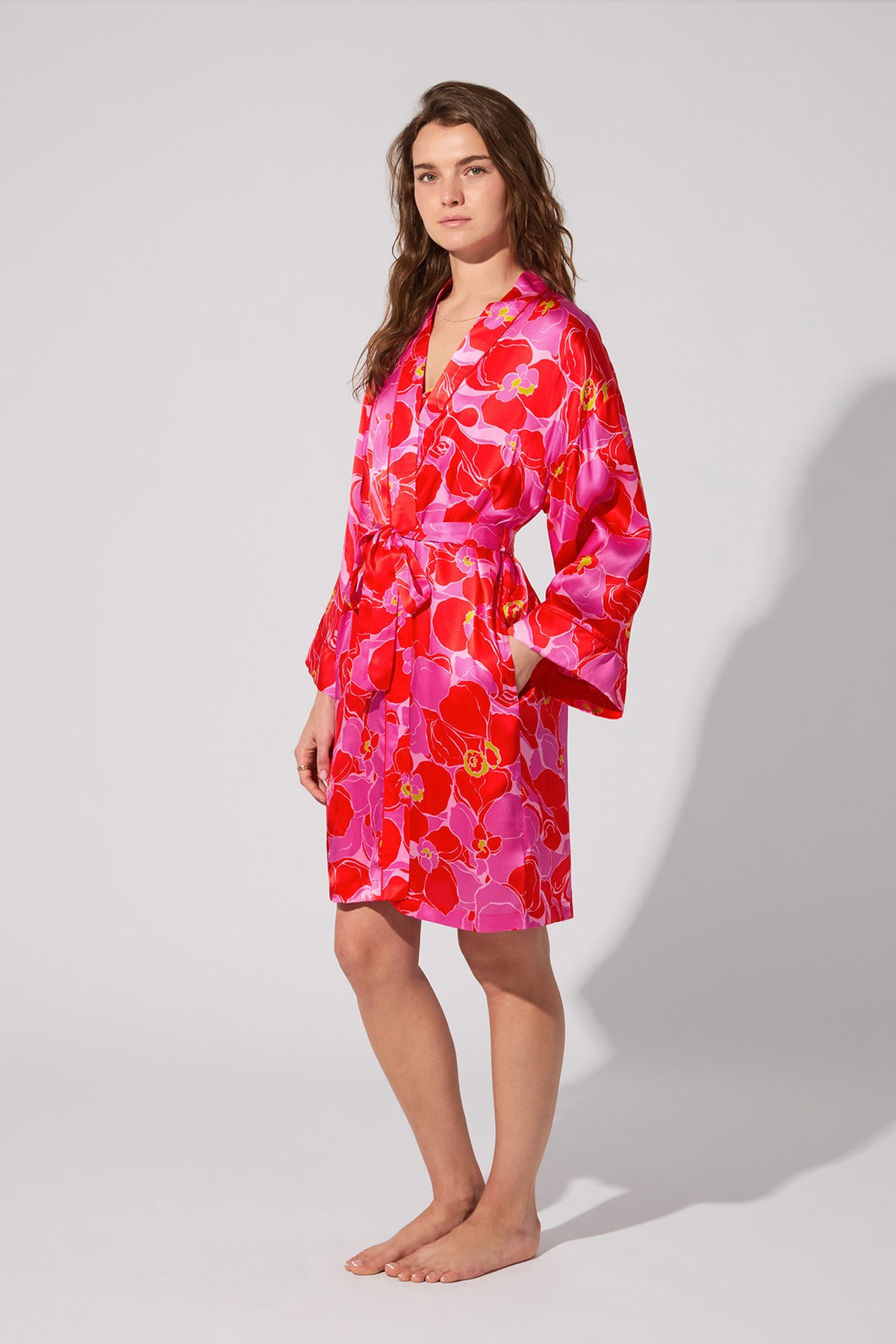 A woman stands barefoot in the Long-Sleeve Silk Robe - Winter Orchids, featuring pink and red floral patterns with a matching belt, looking at the camera against a plain light gray background.