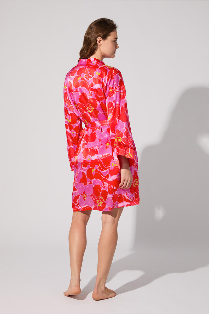 A woman stands barefoot, facing away, wearing the Long-Sleeve Silk Robe - Winter Orchids, featuring pink and red florals with a belted waist, against a plain light background.
