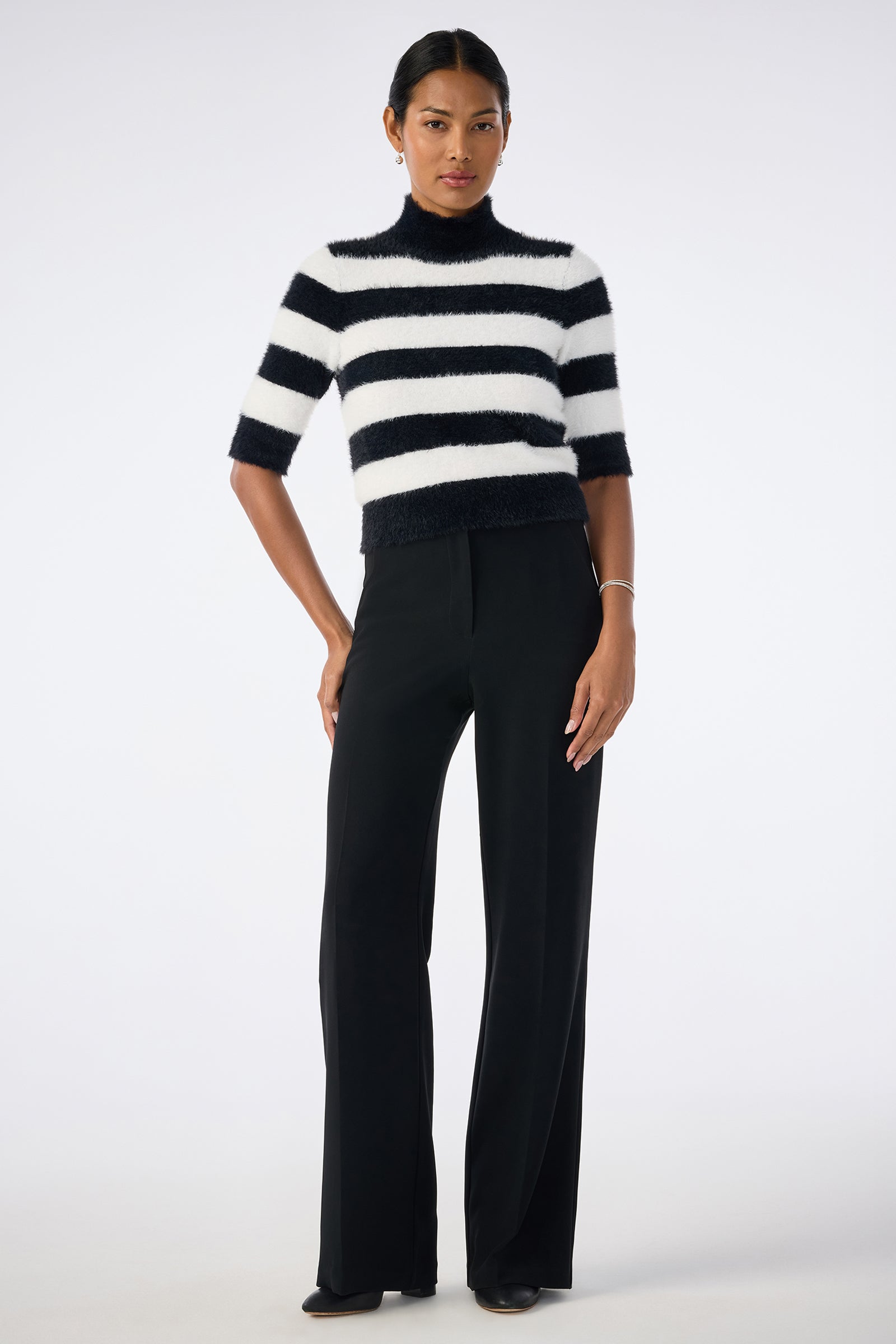 A woman stands facing forward against a plain light background, wearing the Mali Mock Neck Sweater in black and white stripes, paired with black wide-leg trousers and dark shoes. Her hair is pulled back.