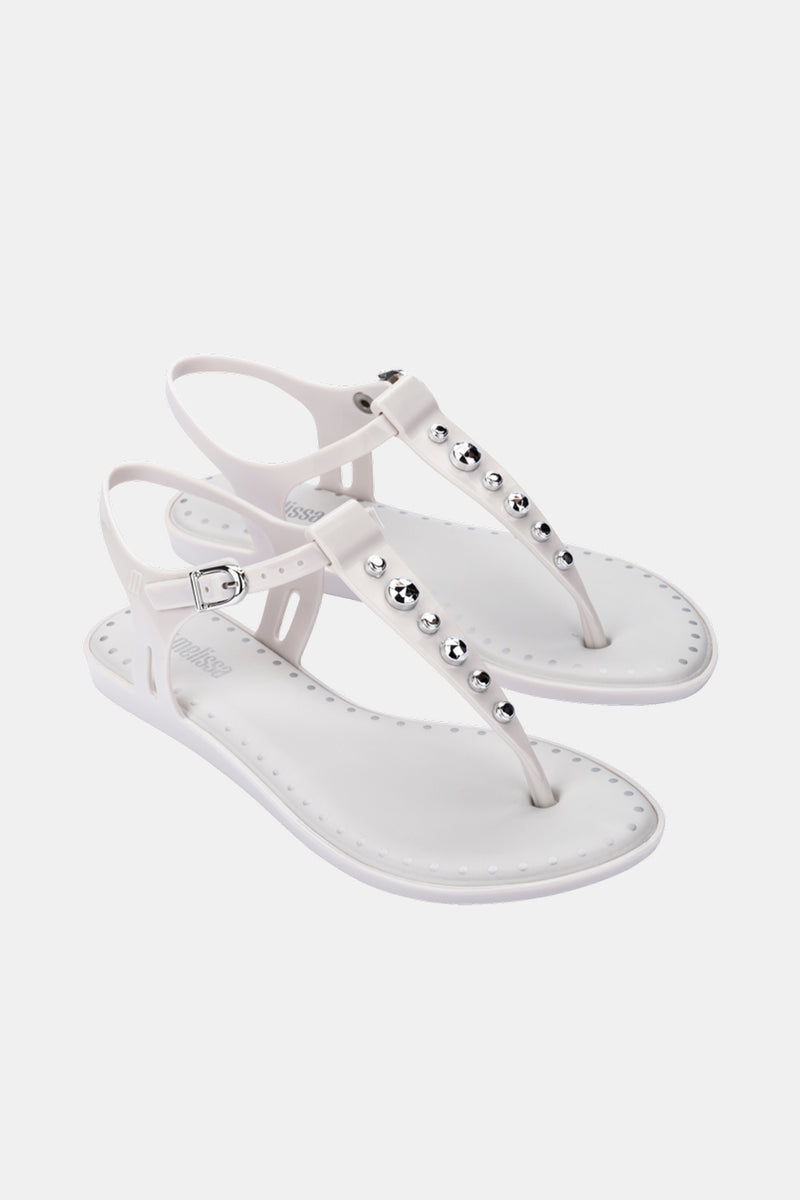 The Melissa Solar Boho is a pair of white flat sandals featuring ankle straps, buckle closure, and silver metallic studs on the T-strap for a minimalist, modern look.