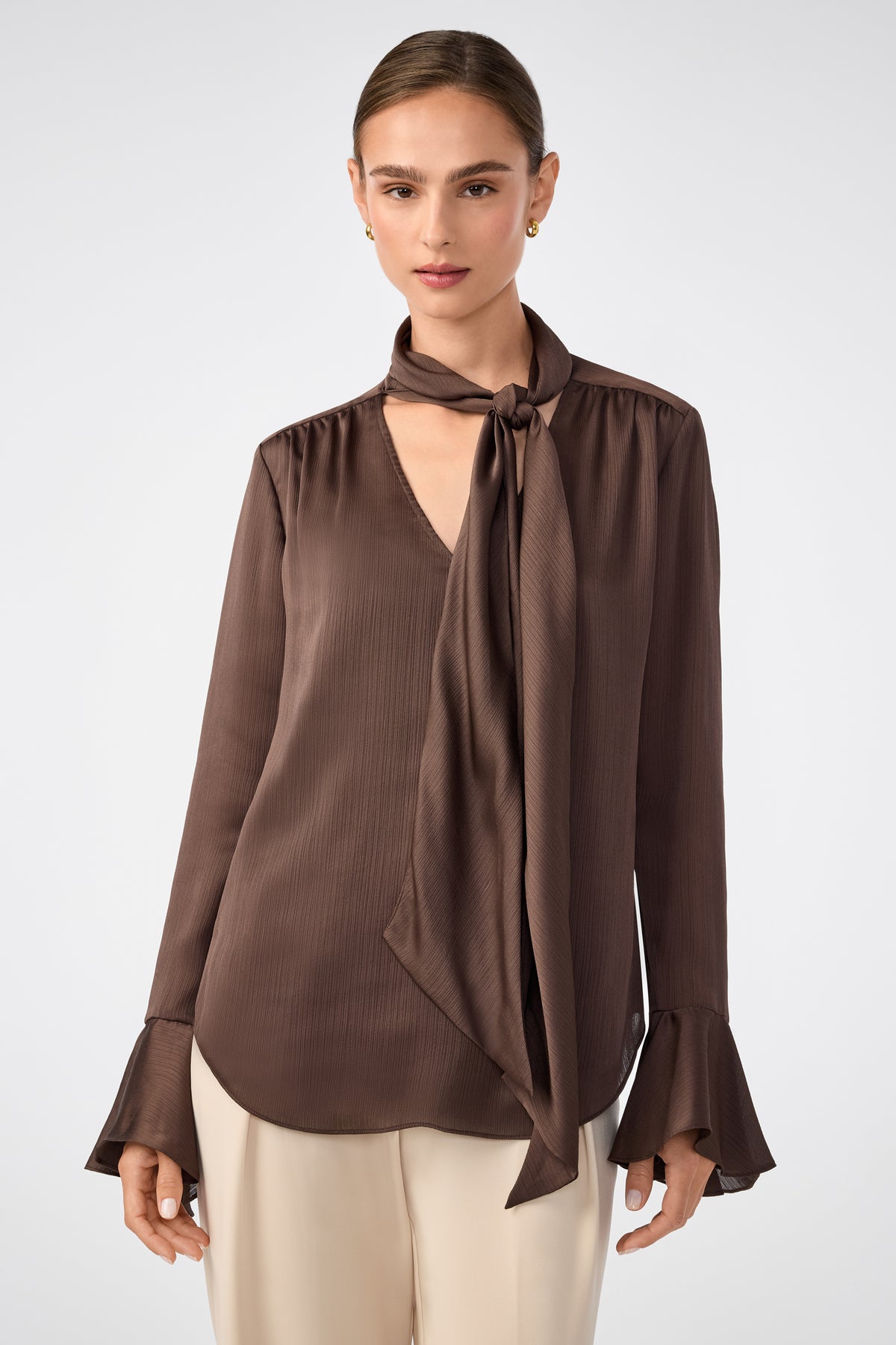 A woman wears the Alora Lavalliere Blouse in walnut, featuring long sleeves, a tie-neck detail, and flared cuffs, paired with light-colored pants, standing against a plain, light background.