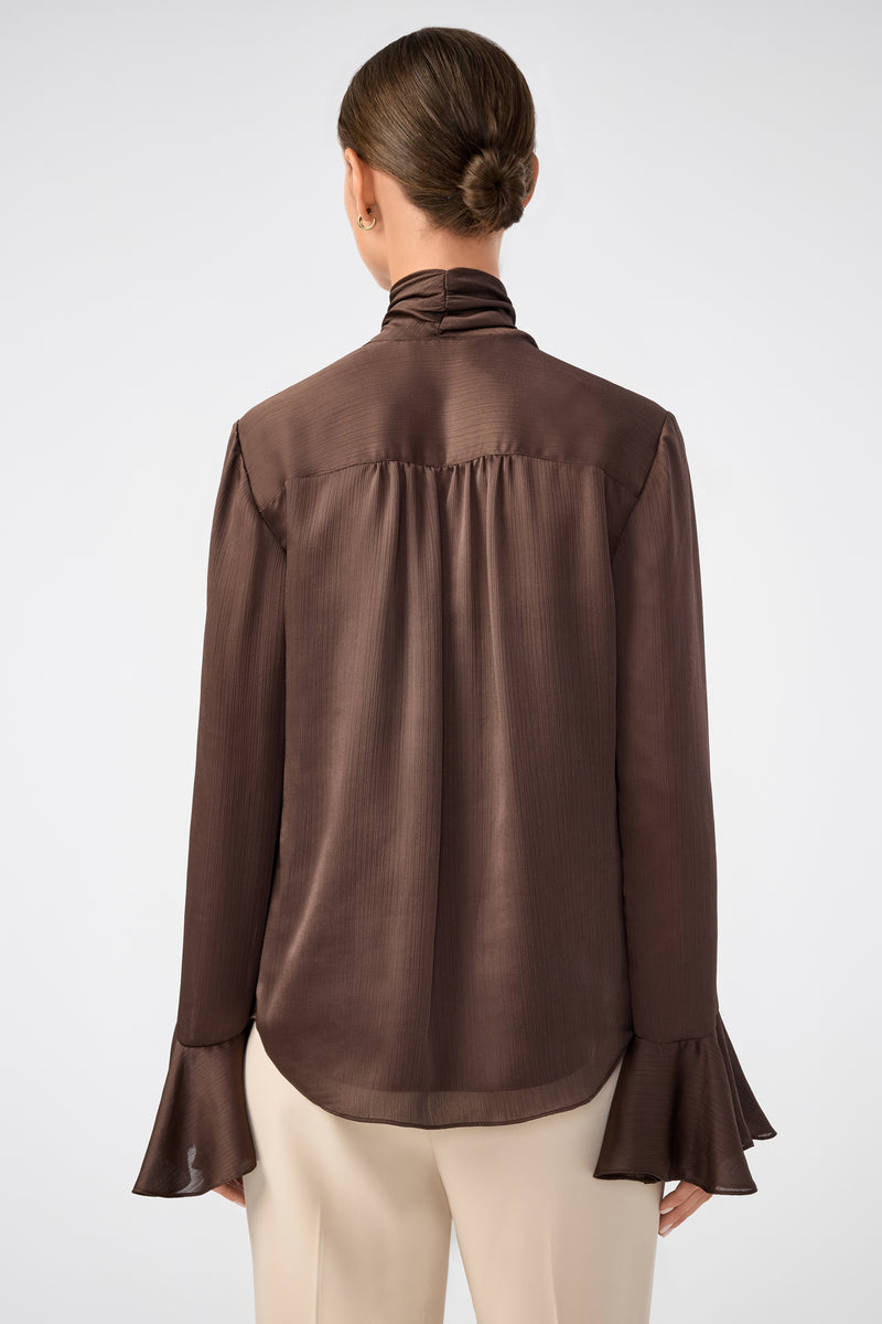 A woman with her hair in a low bun is shown from the back, wearing the Alora Lavalliere Blouse—long-sleeved and dark brown with flared cuffs—paired with cream-colored pants against a plain light background.