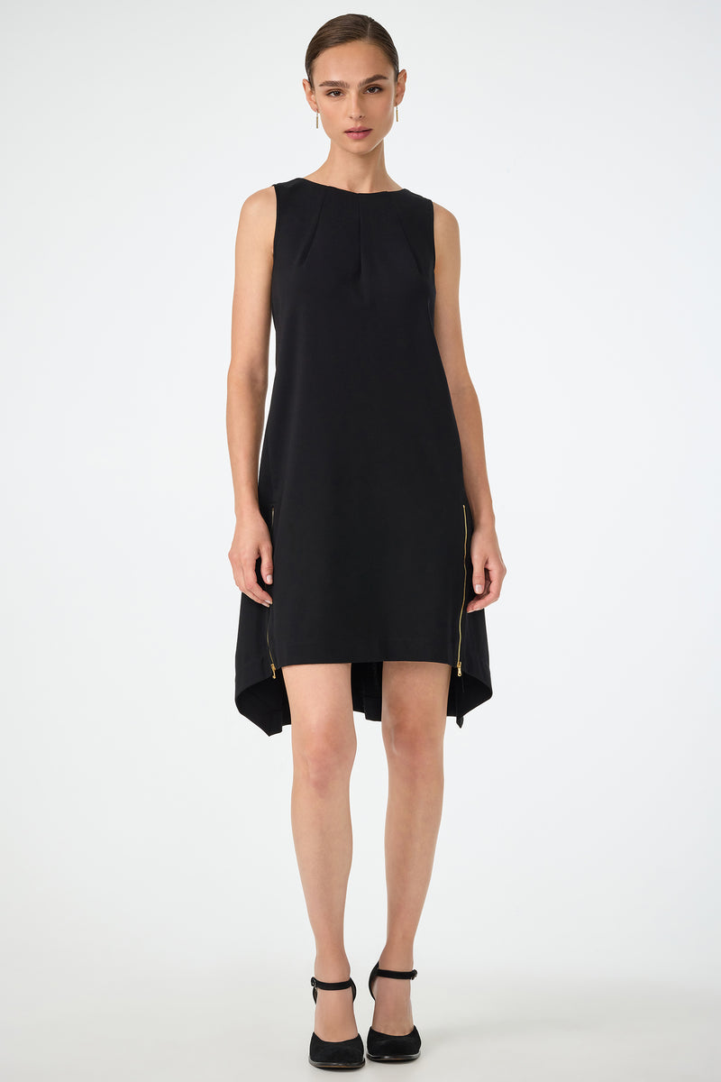 A woman stands facing forward against a white background, wearing the Vantis High-Low Sleeveless Dress in black with pleated neckline detail and gold hip zippers, paired with black ankle-strap heels.
