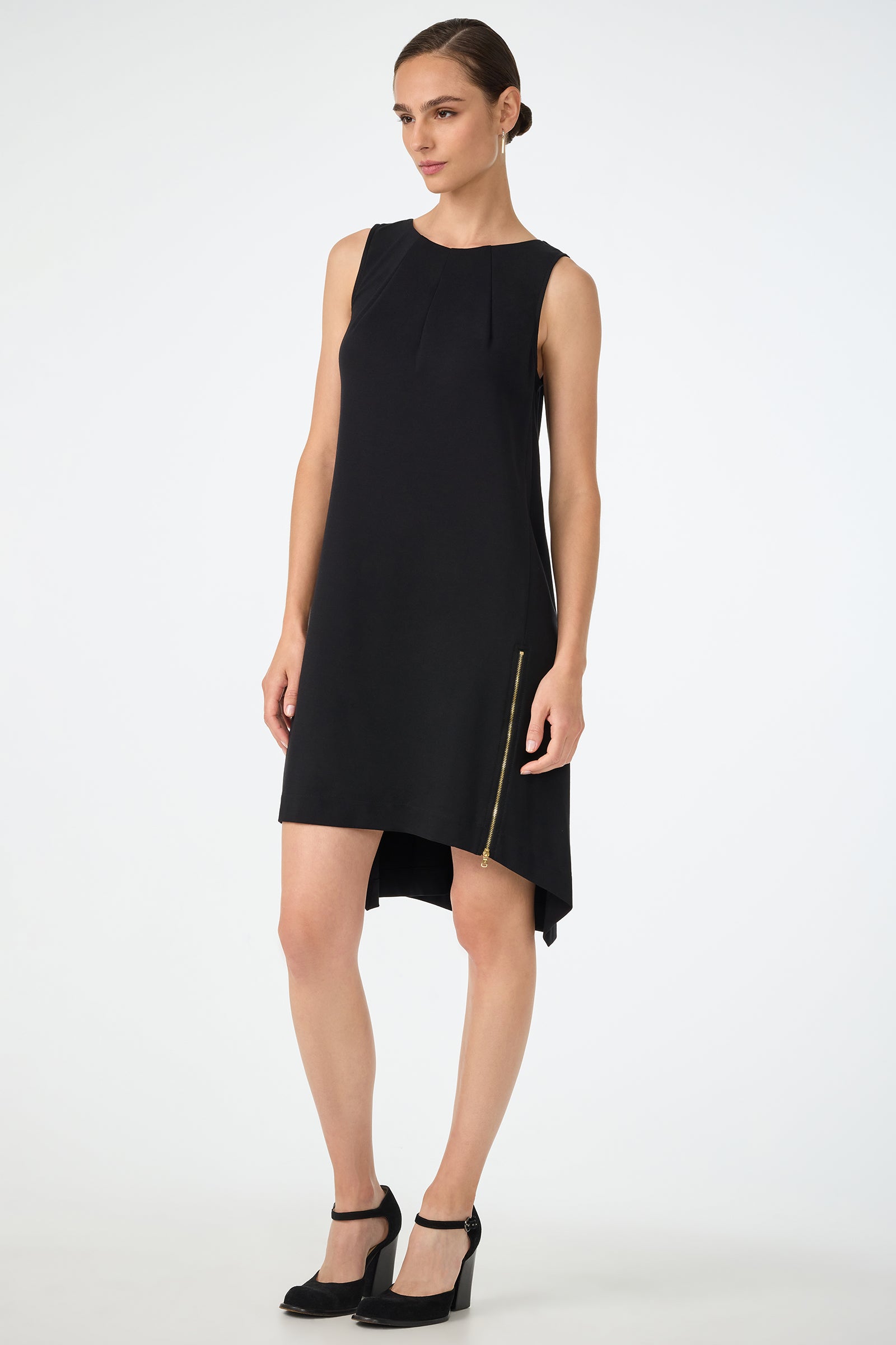 A woman wears the Vantis High-Low Sleeveless Dress, featuring a black asymmetrical hem and gold zipper detail, paired with black heels, standing against a plain white background.