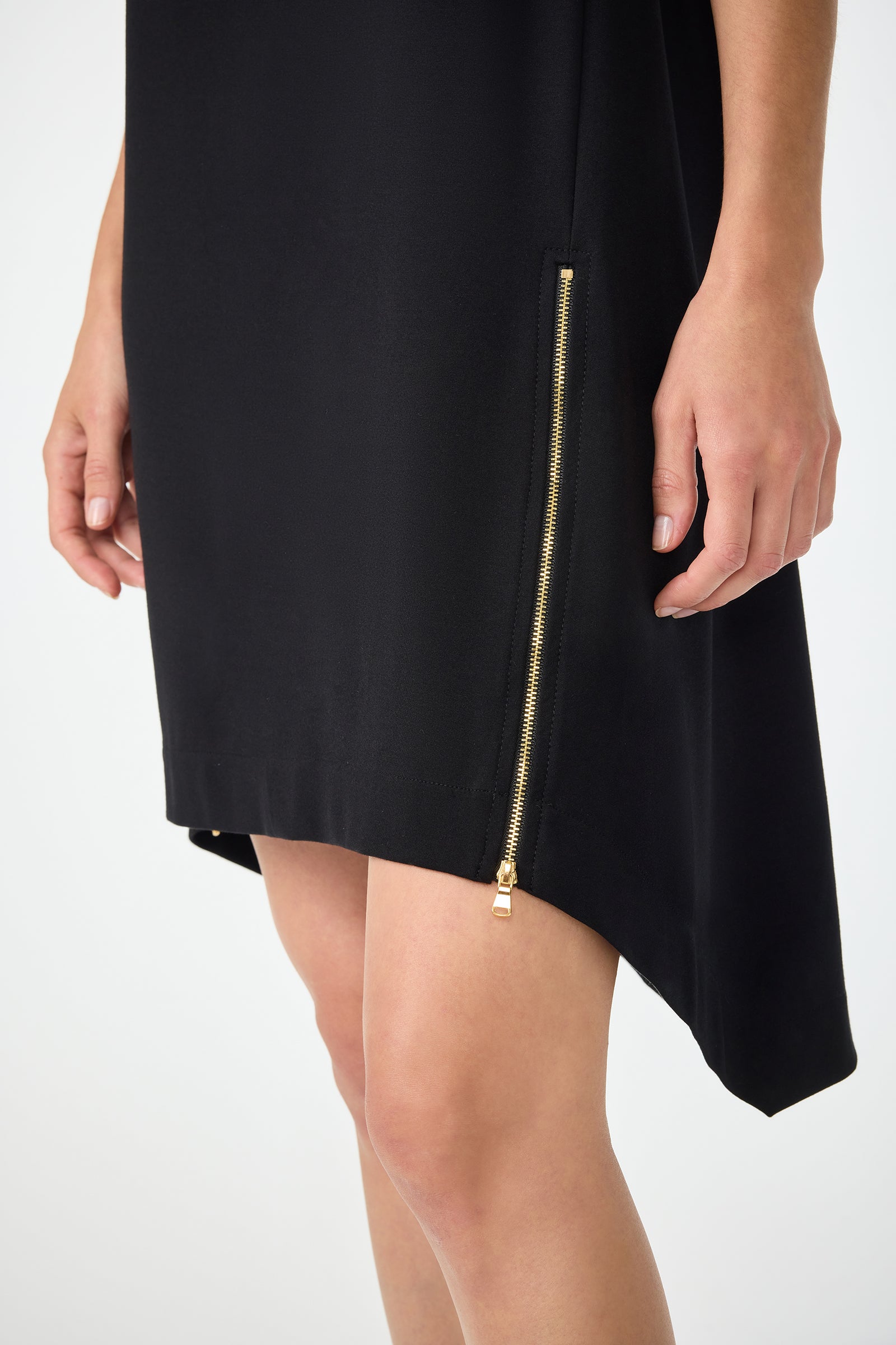 A person wearing the Vantis High-Low Sleeveless Dress in black with a gold zipper detail at the side hem, standing with hands relaxed by their sides. Only the lower half of the dress and upper legs are visible.