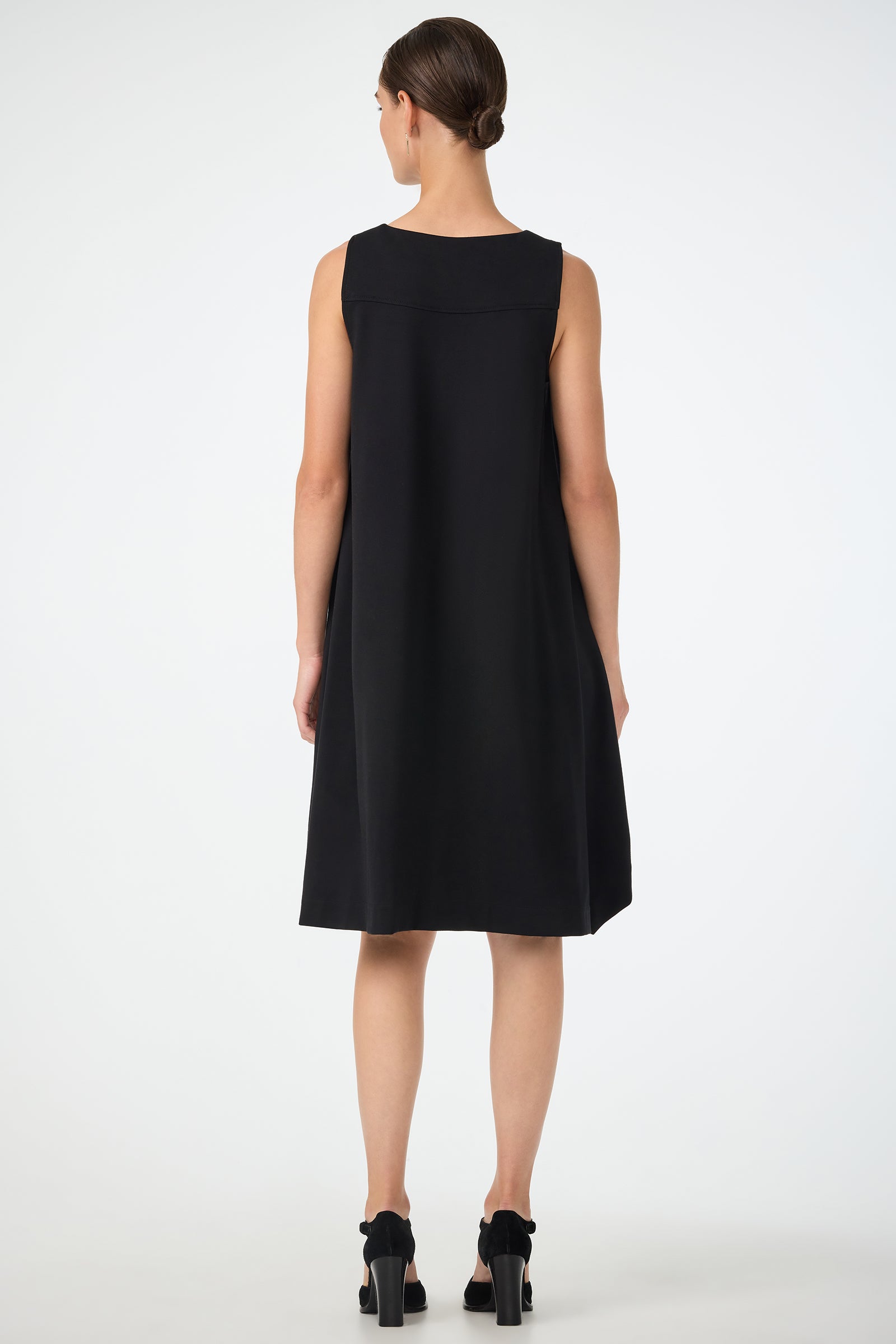 A woman faces away, wearing the Vantis High-Low Sleeveless Dress in black with black heels. Her hair is neatly tied back, and she stands against a plain white background.