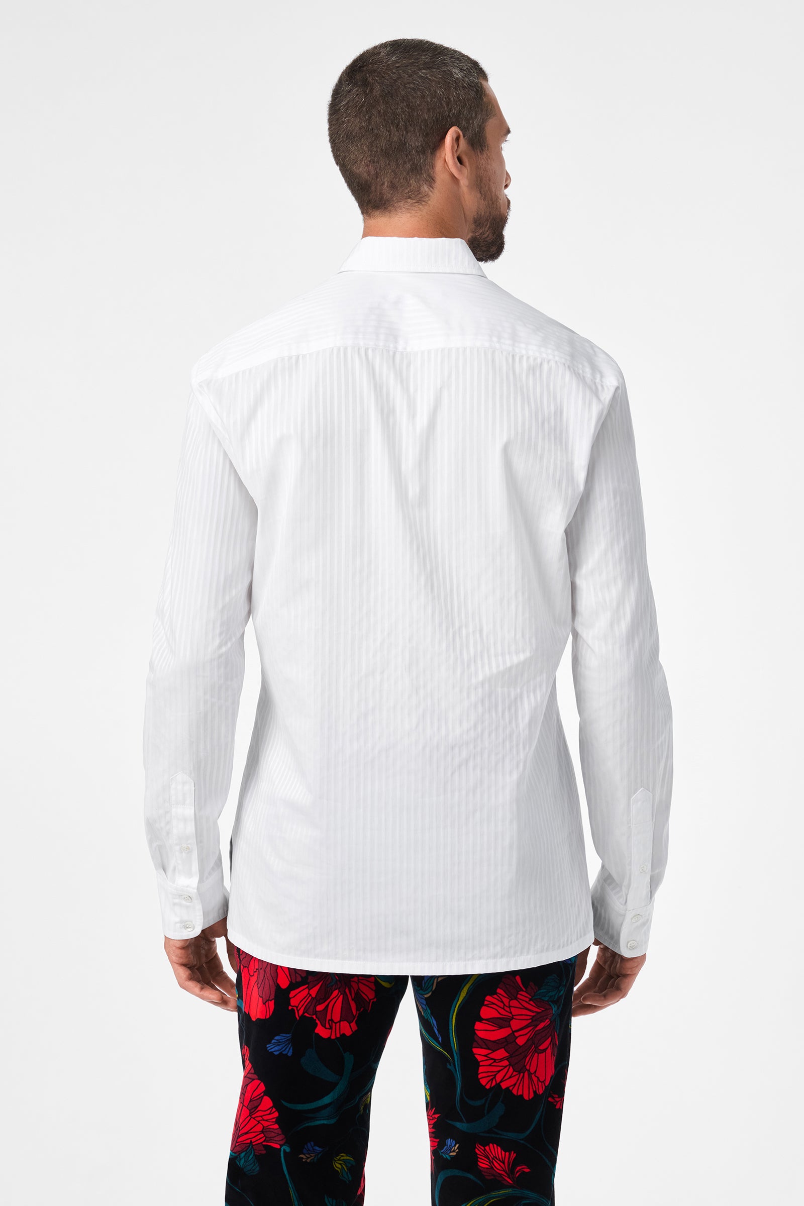 A man stands with his back to the camera, wearing the Evan Tux Shirt—white, long-sleeved—and black pants with a striking red and blue floral pattern. The background is plain white.
