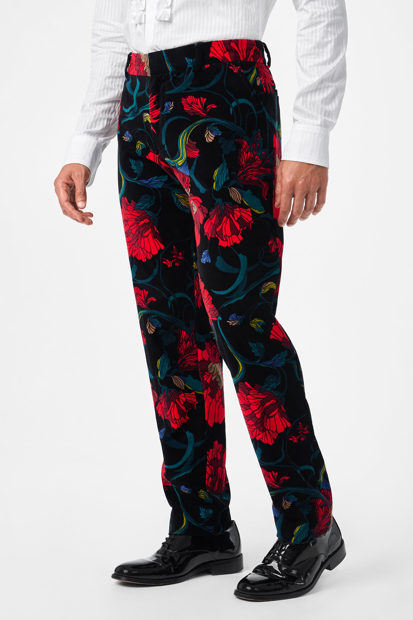 A person wearing the Clyde Slim Printed Velvet Trouser featuring a bold red and blue floral pattern, paired with a white dress shirt and black shiny dress shoes, stands against a plain background.