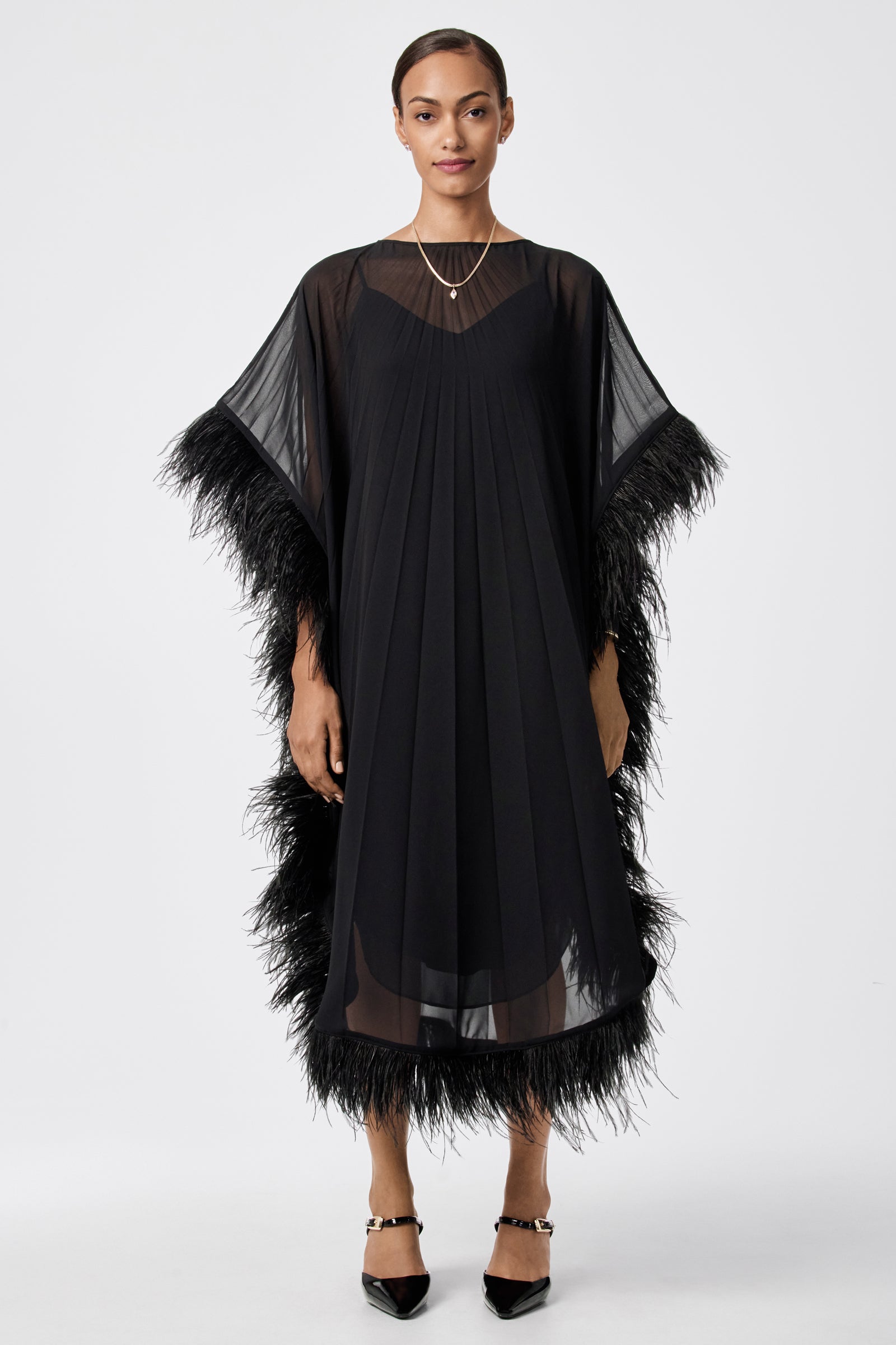 A woman stands facing forward in the Pleated Neena Feather-Trimmed Caftan Dress, featuring sheer sleeves and feather trim. She has short hair, minimal jewelry, and wears black pointed shoes against a plain white background.