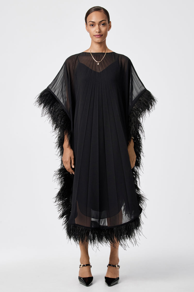 A woman stands facing forward in the Pleated Neena Feather-Trimmed Caftan Dress, featuring sheer sleeves and feather trim. She has short hair, minimal jewelry, and wears black pointed shoes against a plain white background.