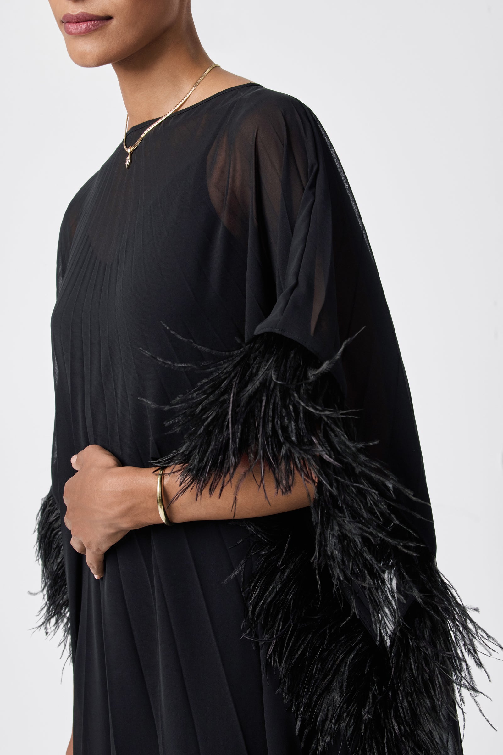 A person wearing the Pleated Neena Feather-Trimmed Caftan Dress with sheer fabric and black feather-trimmed sleeves, styled with a gold bracelet and delicate gold necklace, stands against a plain white background.